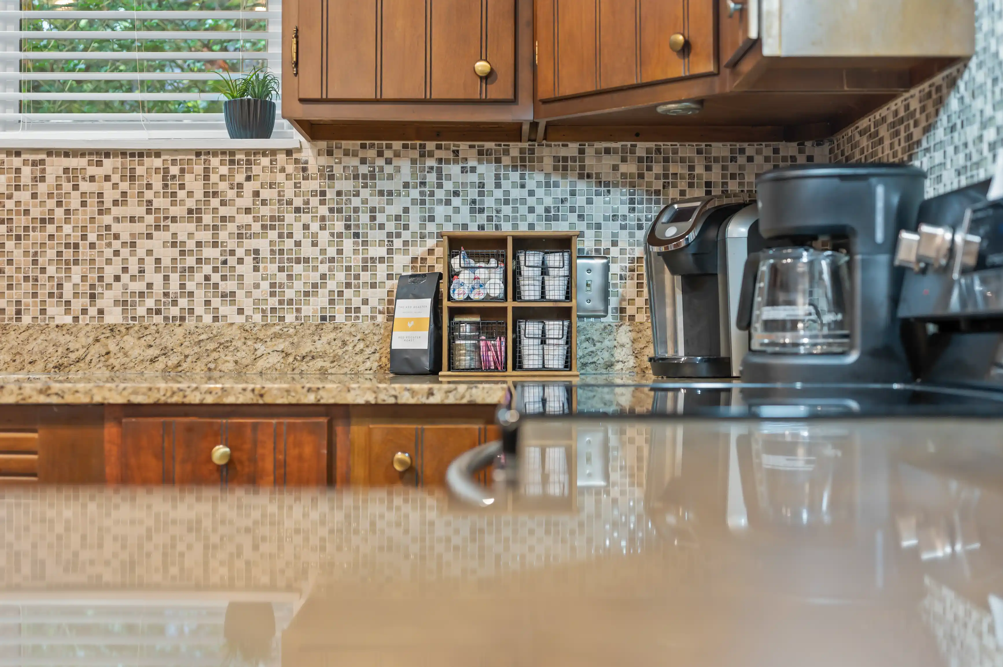 Modern kitchen countertop with reflective surface, showing a coffee maker, a wooden organizer with coffee pods, and a potted plant near the window with mosaic tile backsplash.