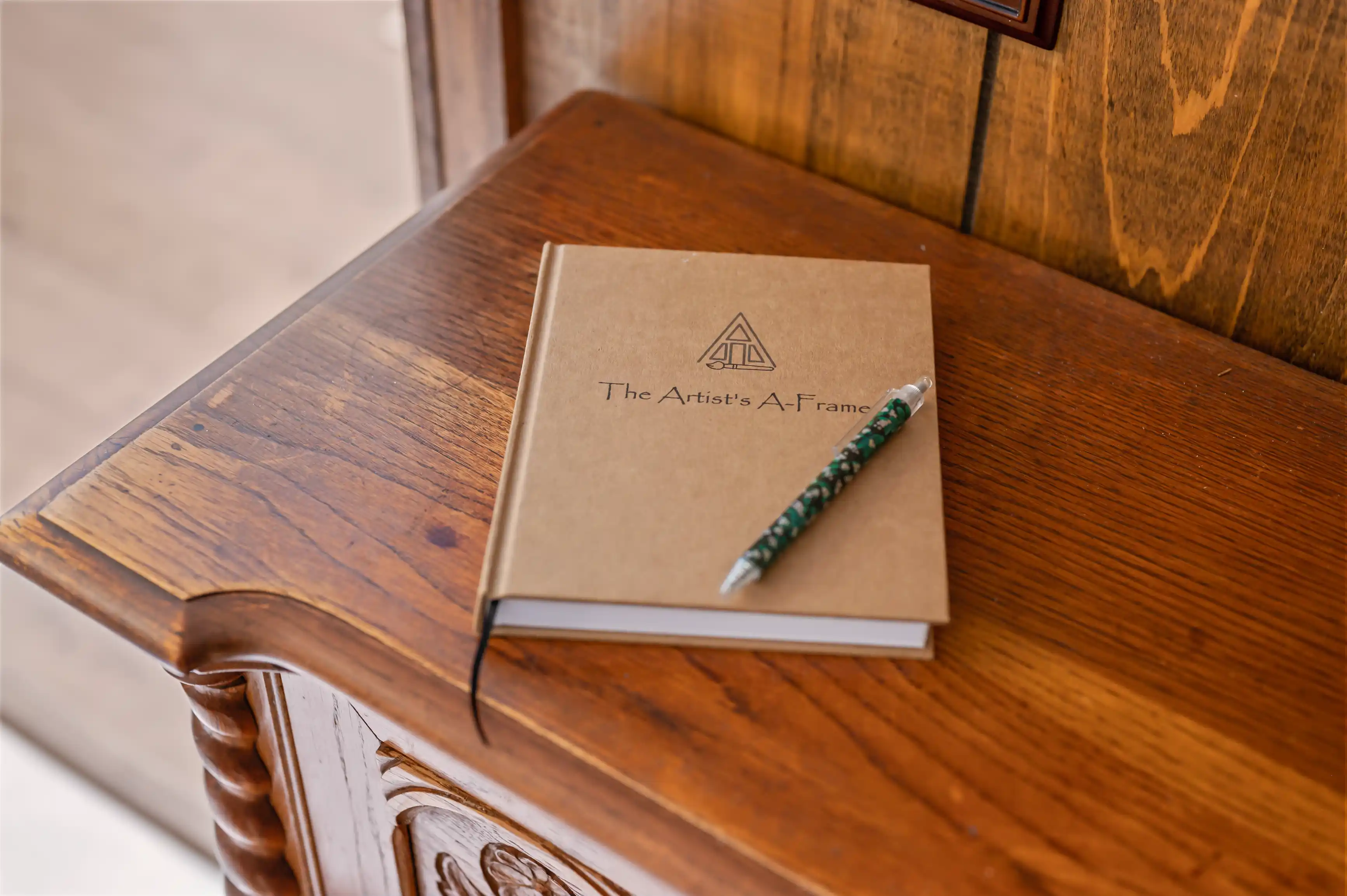 A closed book titled "The Artist's A-Frame" with a logo on the cover next to a patterned pencil, resting on a wooden surface beside an ornate piece of furniture.