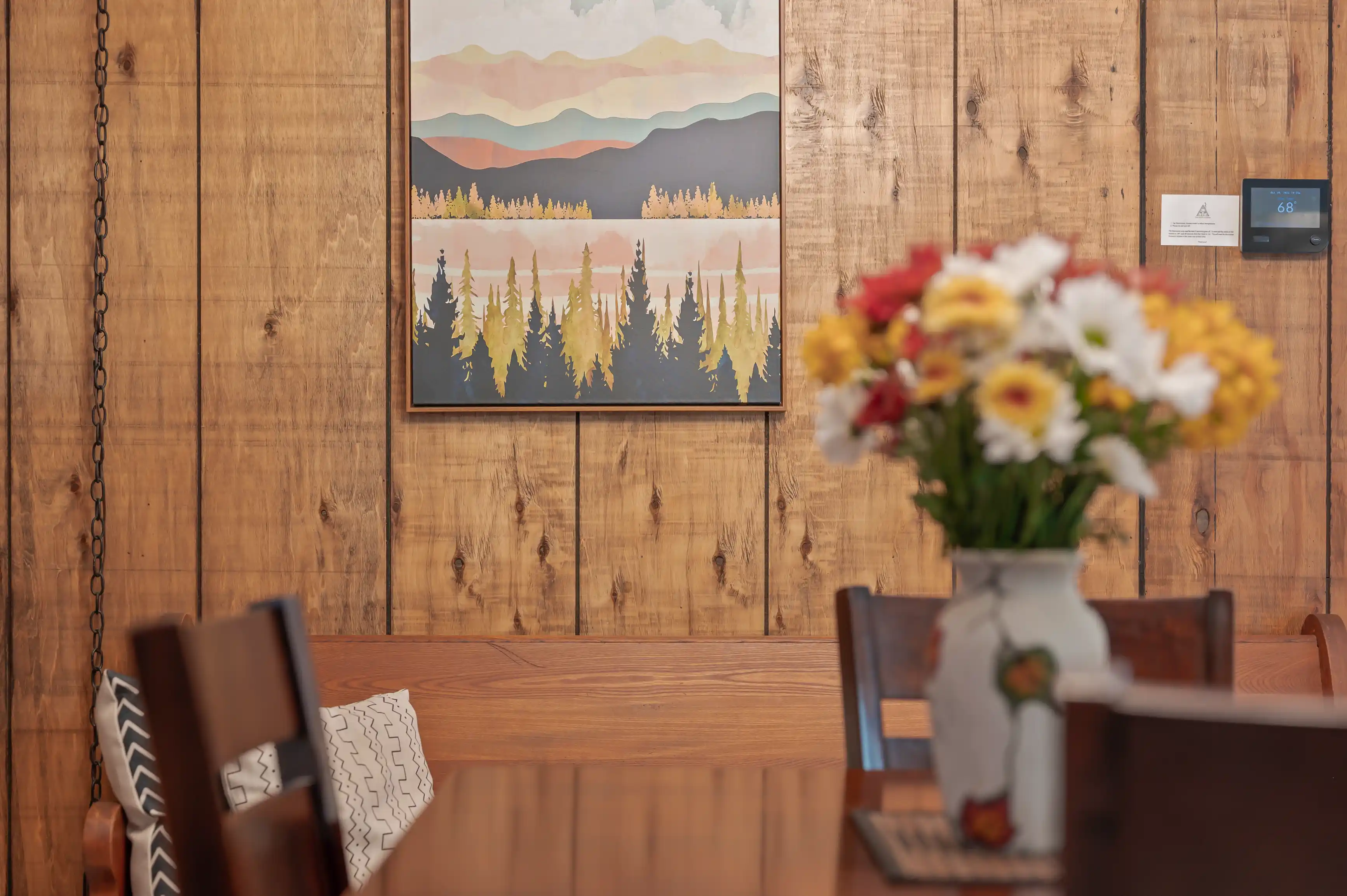 An interior view of a cozy room with wooden walls, a landscape painting hanging on the wall, a wooden table with chairs, and a vase of colorful flowers in the foreground.