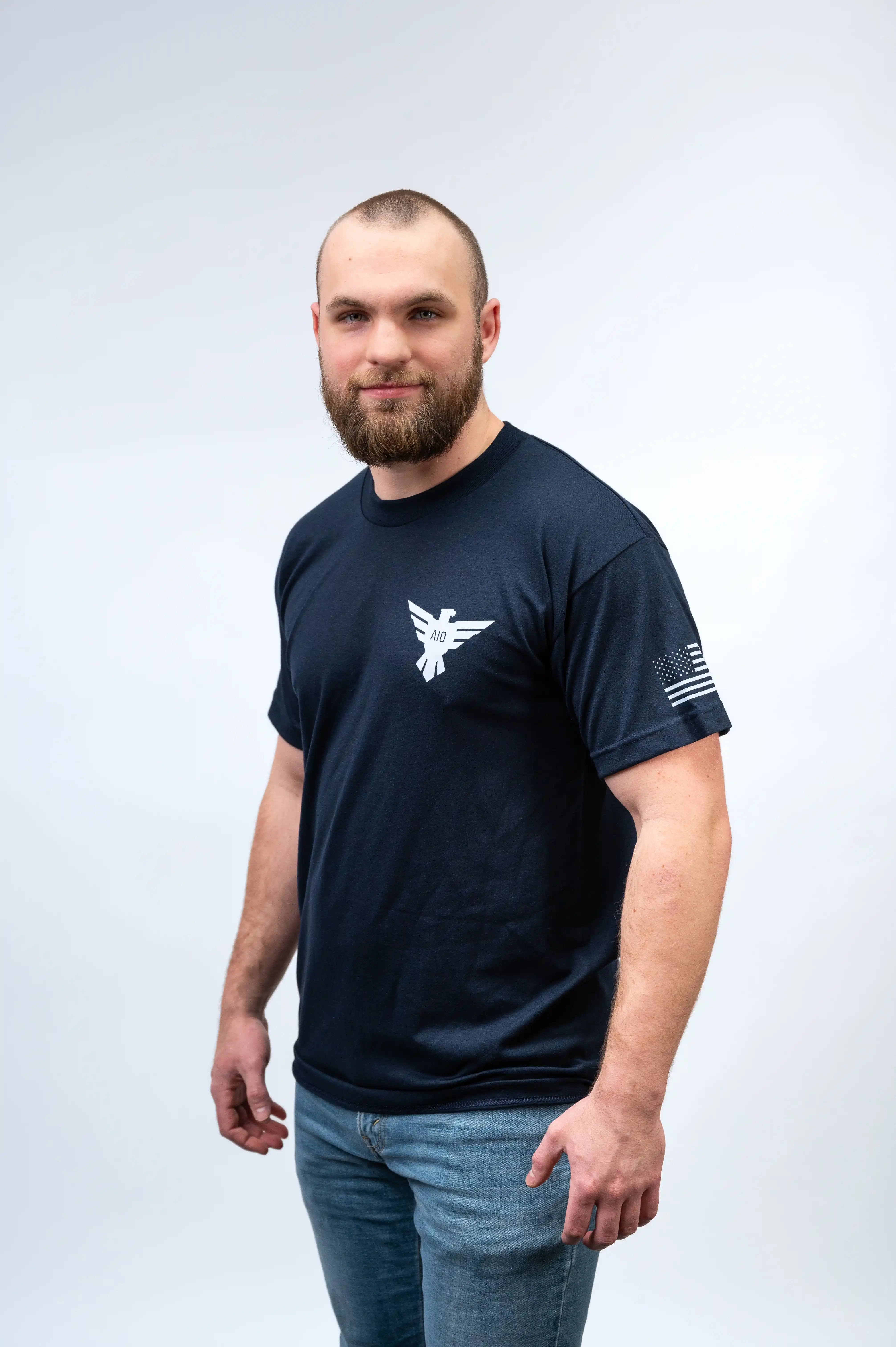 Man with a beard in a navy blue t-shirt with a logo on the chest and an American flag on the right sleeve, standing against a light background.