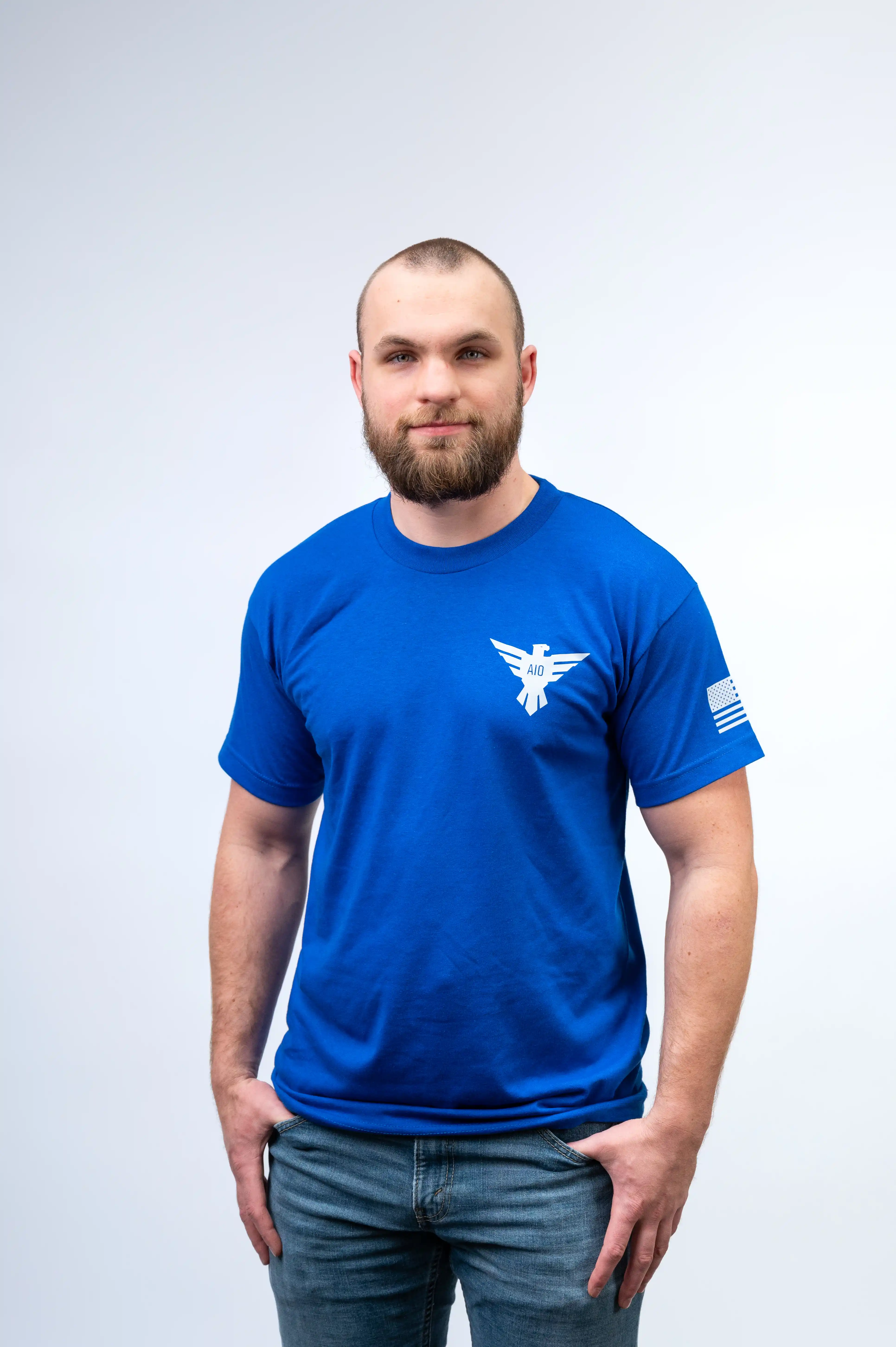 Man with a beard wearing a blue t-shirt standing against a light background.
