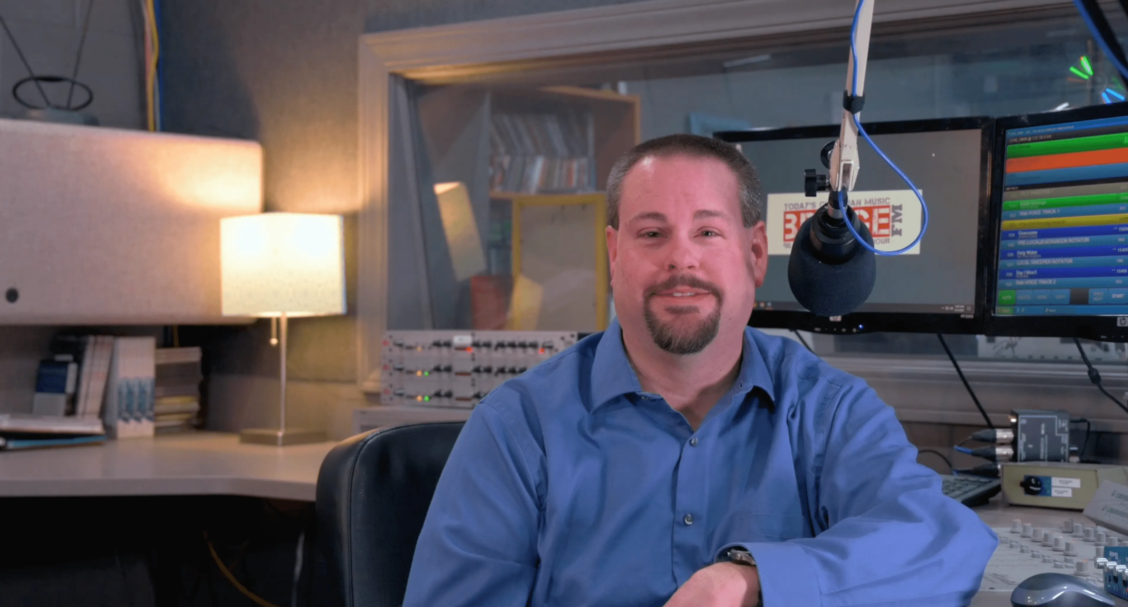 Man in blue shirt smiling at camera in a radio station studio with microphone and control board equipment in the background.