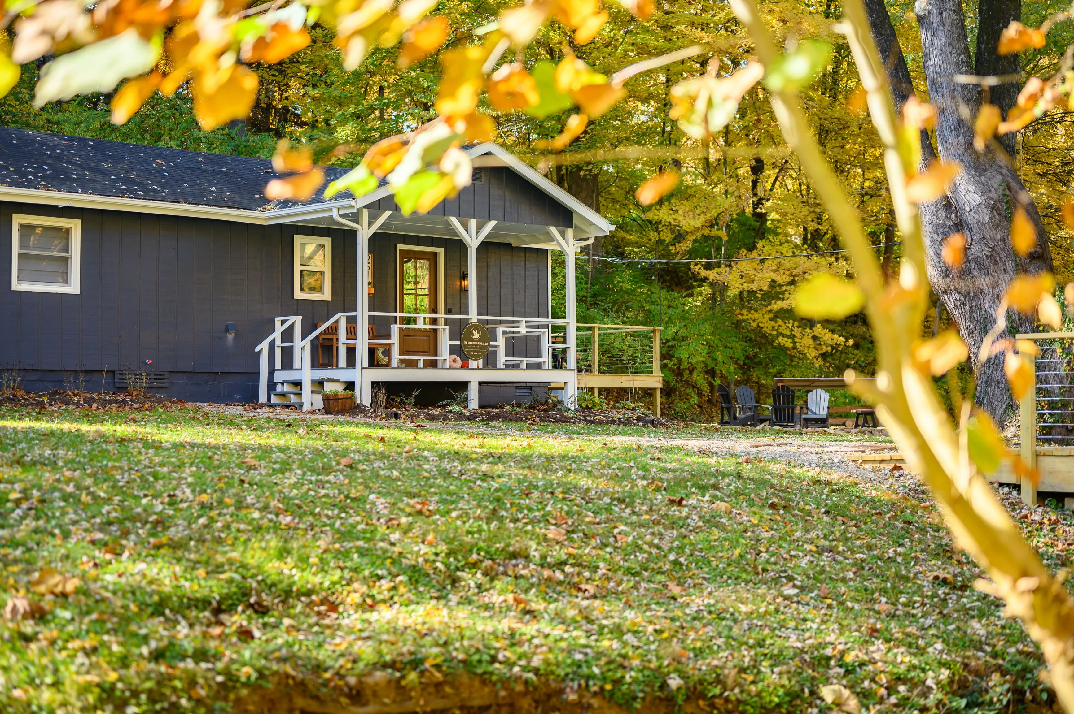 Cozy blue house with white trim and front porch surrounded by autumn foliage and a lawn covered with fallen leaves.