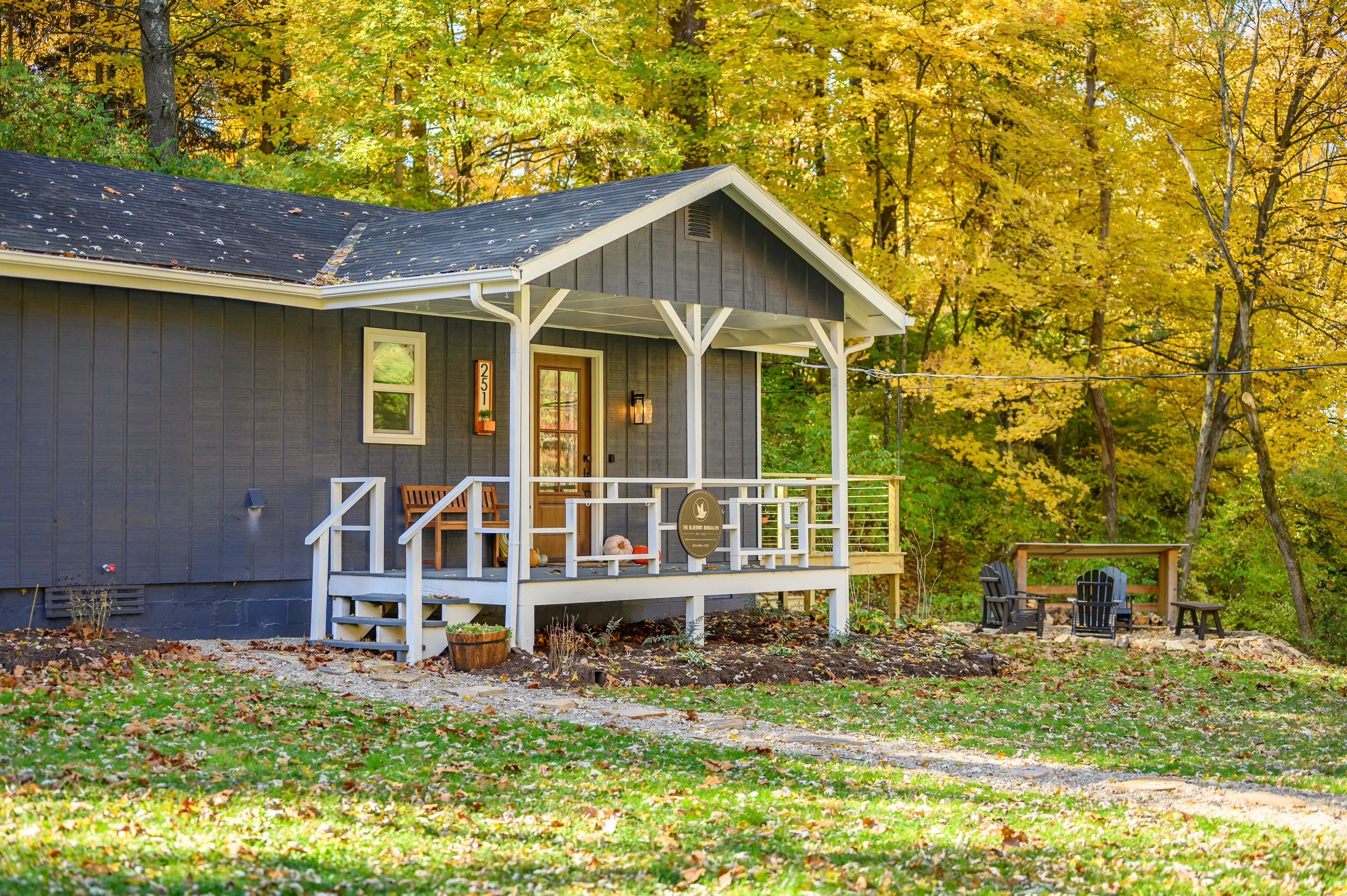 A cozy gray cabin with a front porch surrounded by autumn foliage with a bench and chairs outside under the trees.