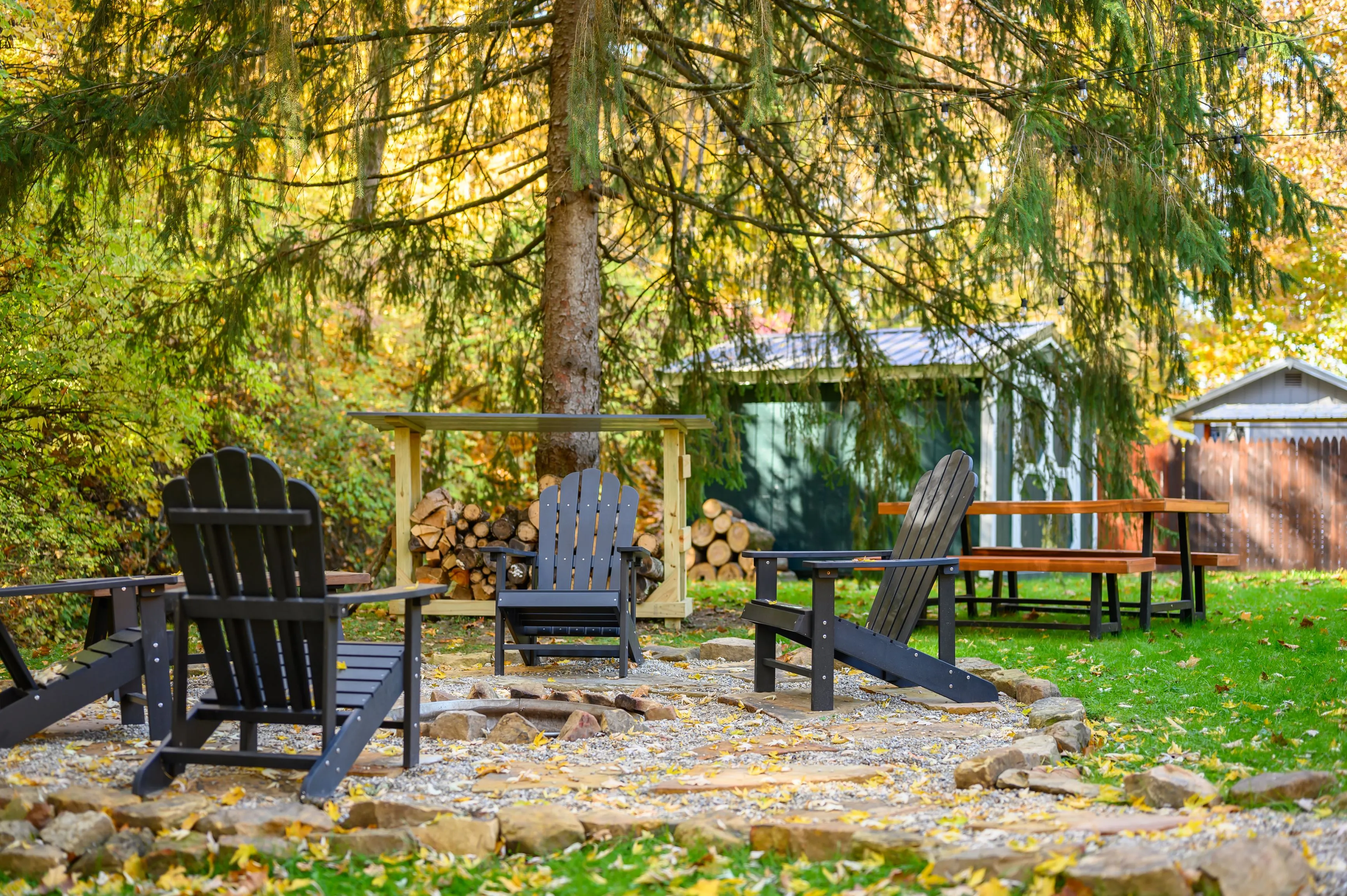A peaceful outdoor seating area with Adirondack chairs around a fire pit, set against a backdrop of autumn foliage and a rustic wooden shed.
