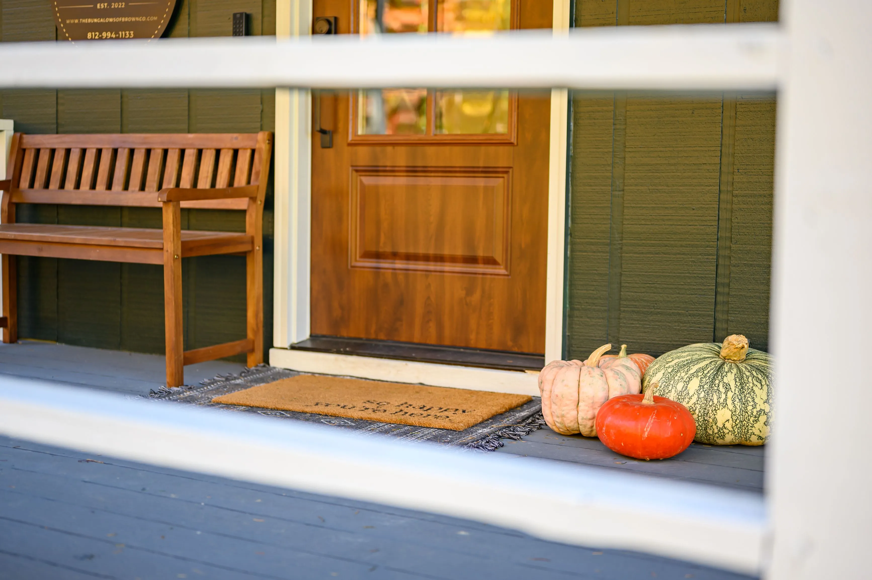 A cozy front porch view with a wooden bench next to a door, decorated with a variety of pumpkins on the ground.