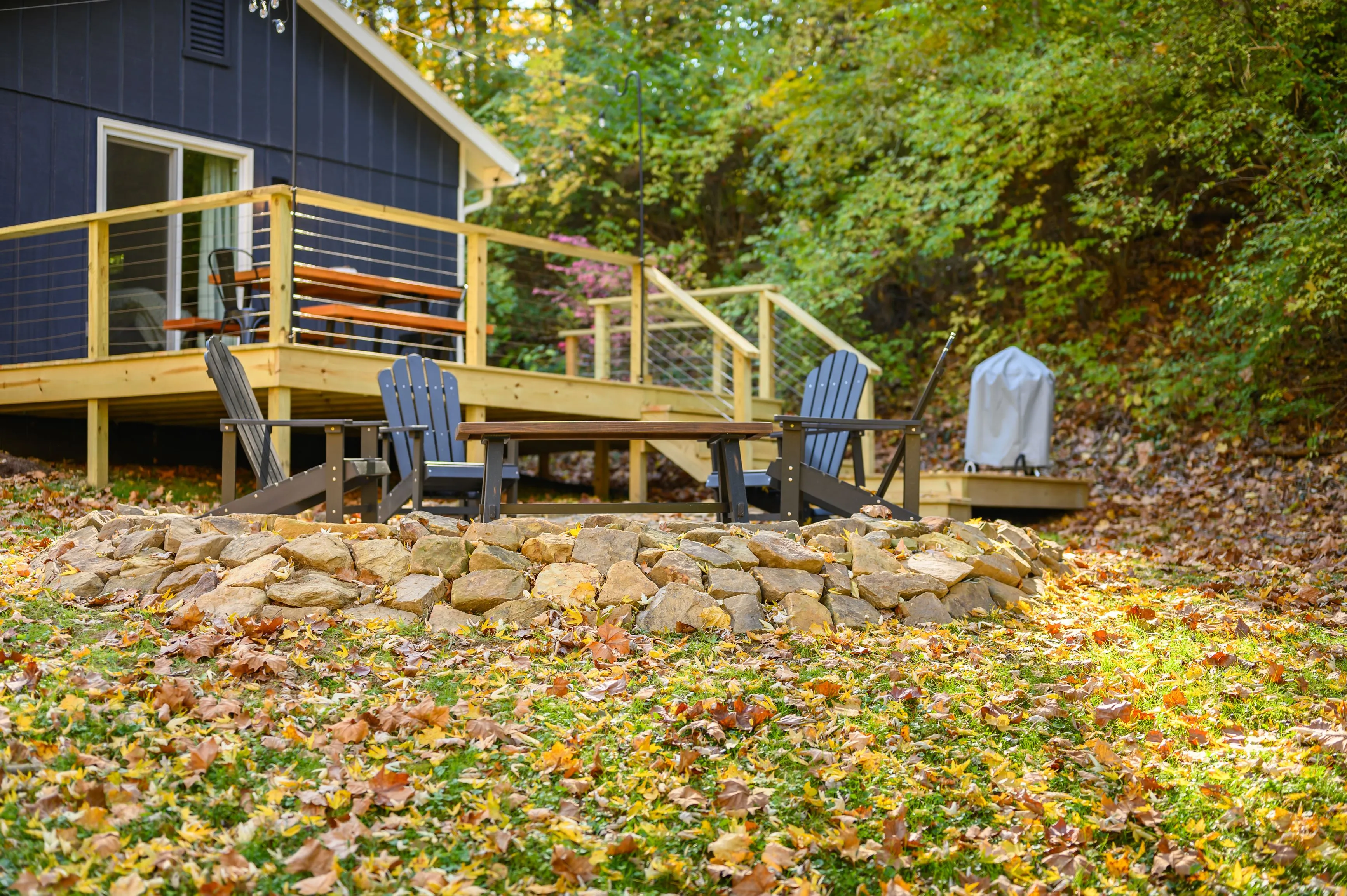 Cozy backyard deck with Adirondack chairs and a stone fire pit surrounded by autumn leaves.