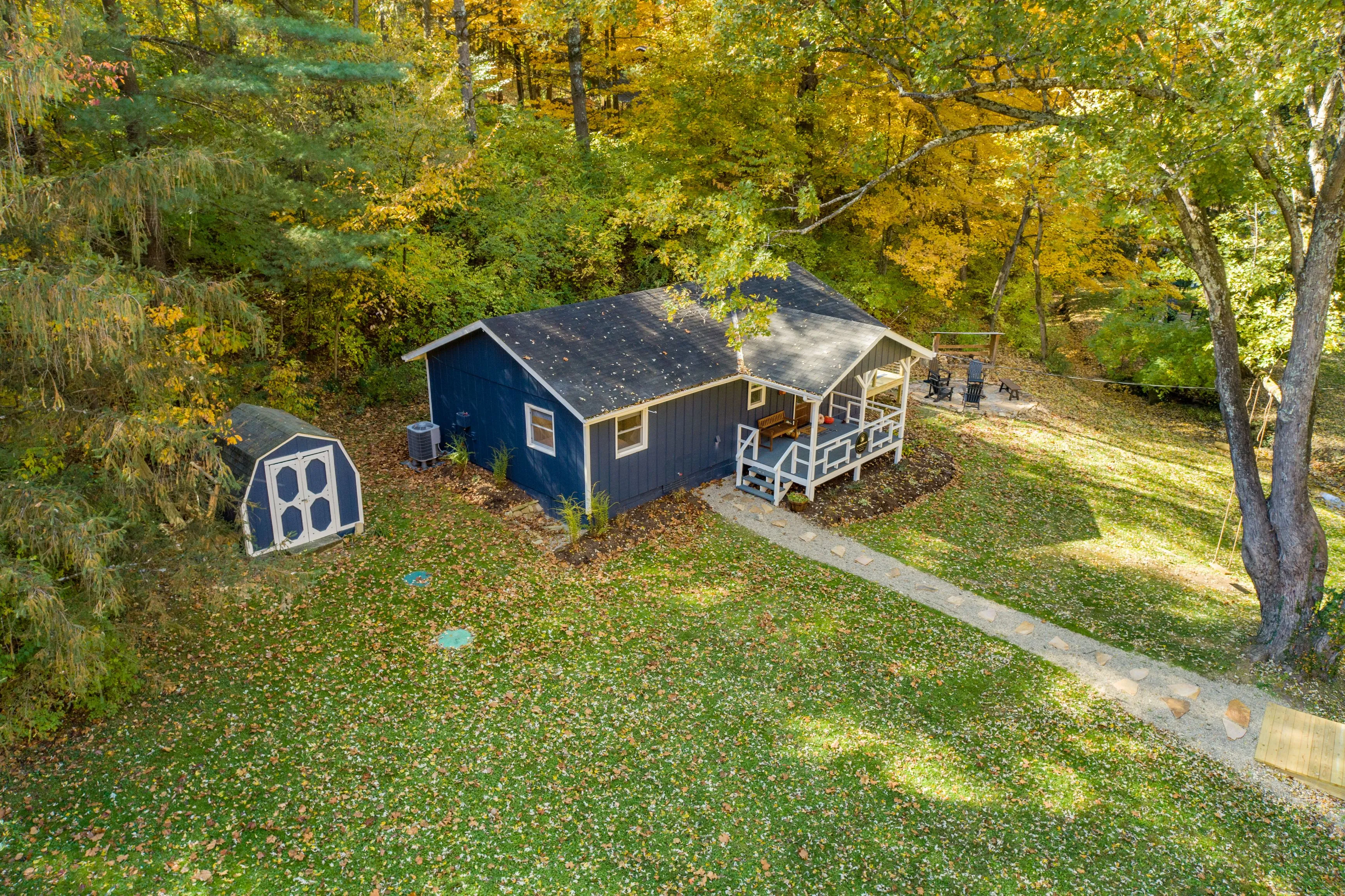 Aerial view of a small blue house with a white-trimmed porch surrounded by autumnal trees with a garden shed and fire pit in the yard.
