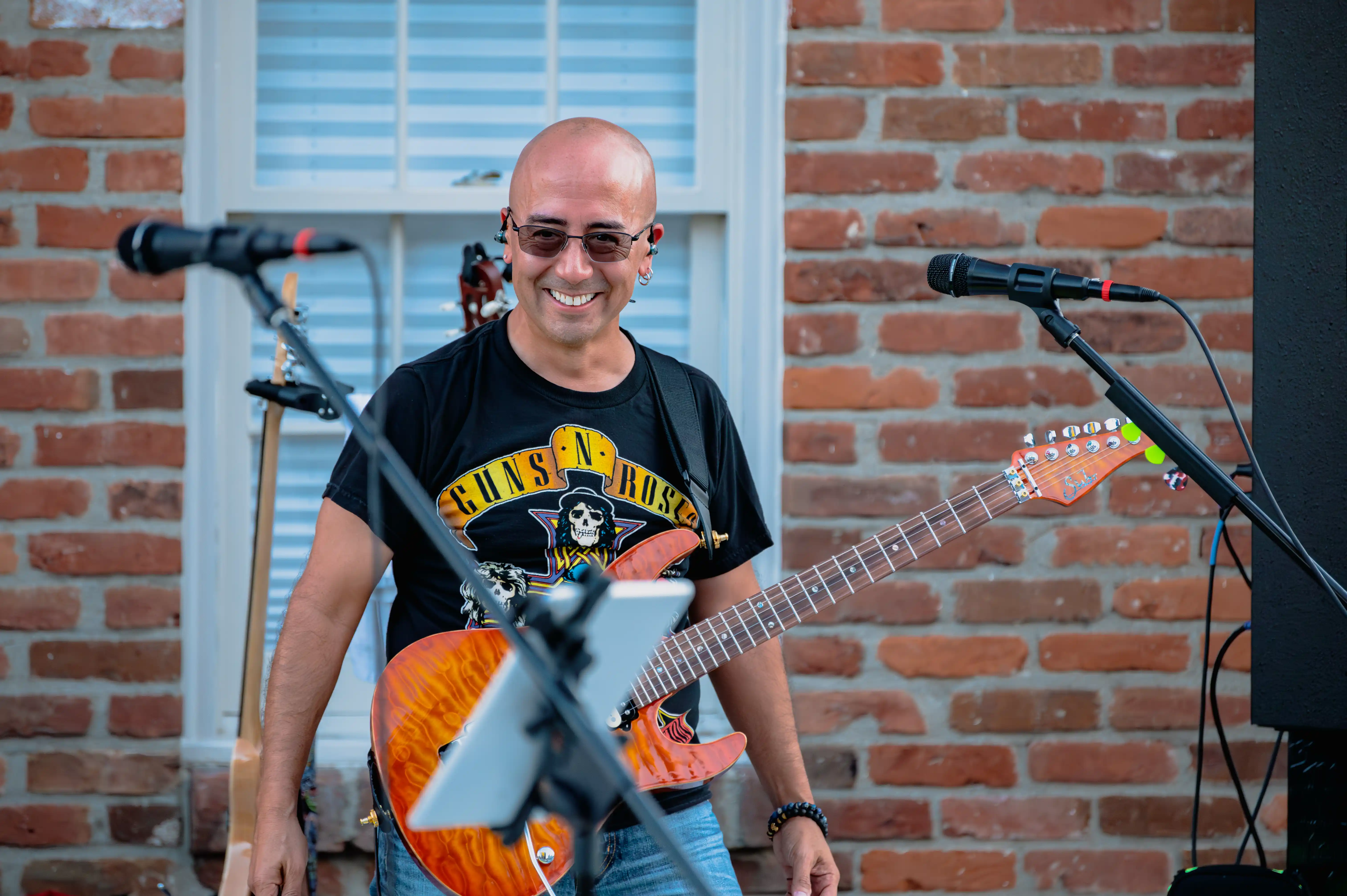 Smiling musician with sunglasses playing an orange electric guitar in front of a microphone and a brick wall background.