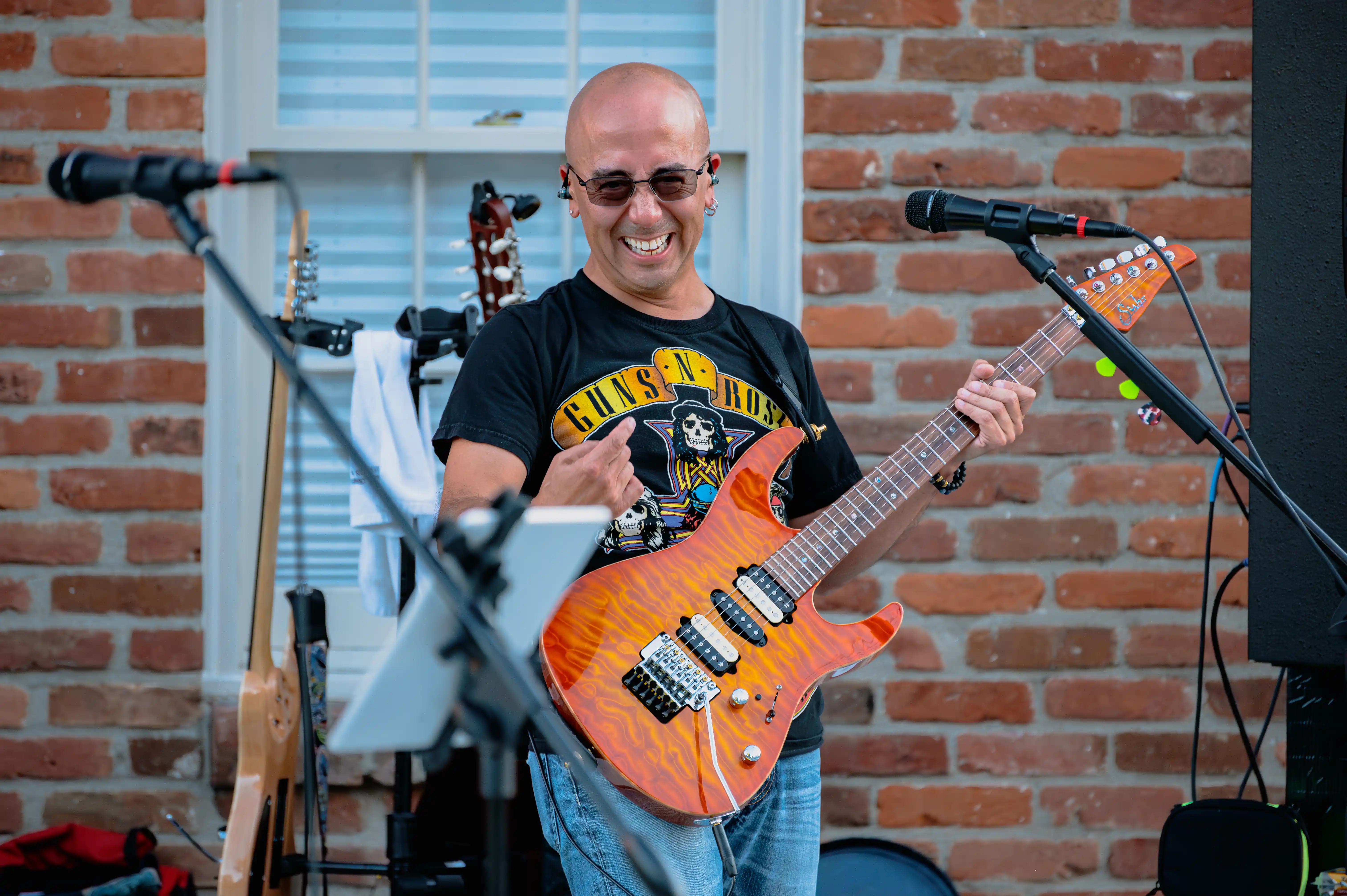 Smiling musician playing an electric guitar on stage with microphone stands and a brick wall in the background.