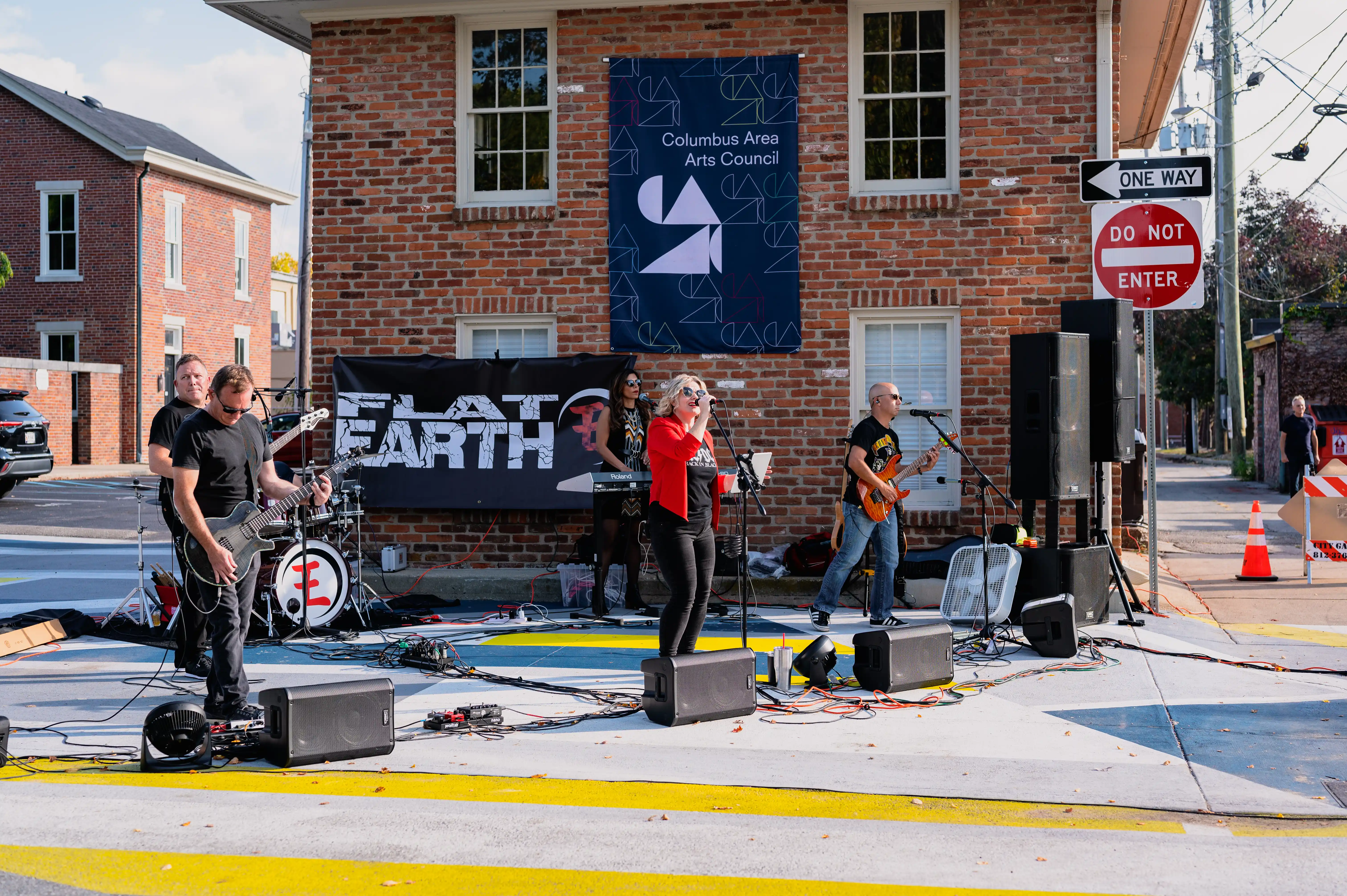 A street band performs outdoors with a drummer, guitarist, and vocalist, a banner reading "Flat Earth" in the background, set against a brick building and street signs.