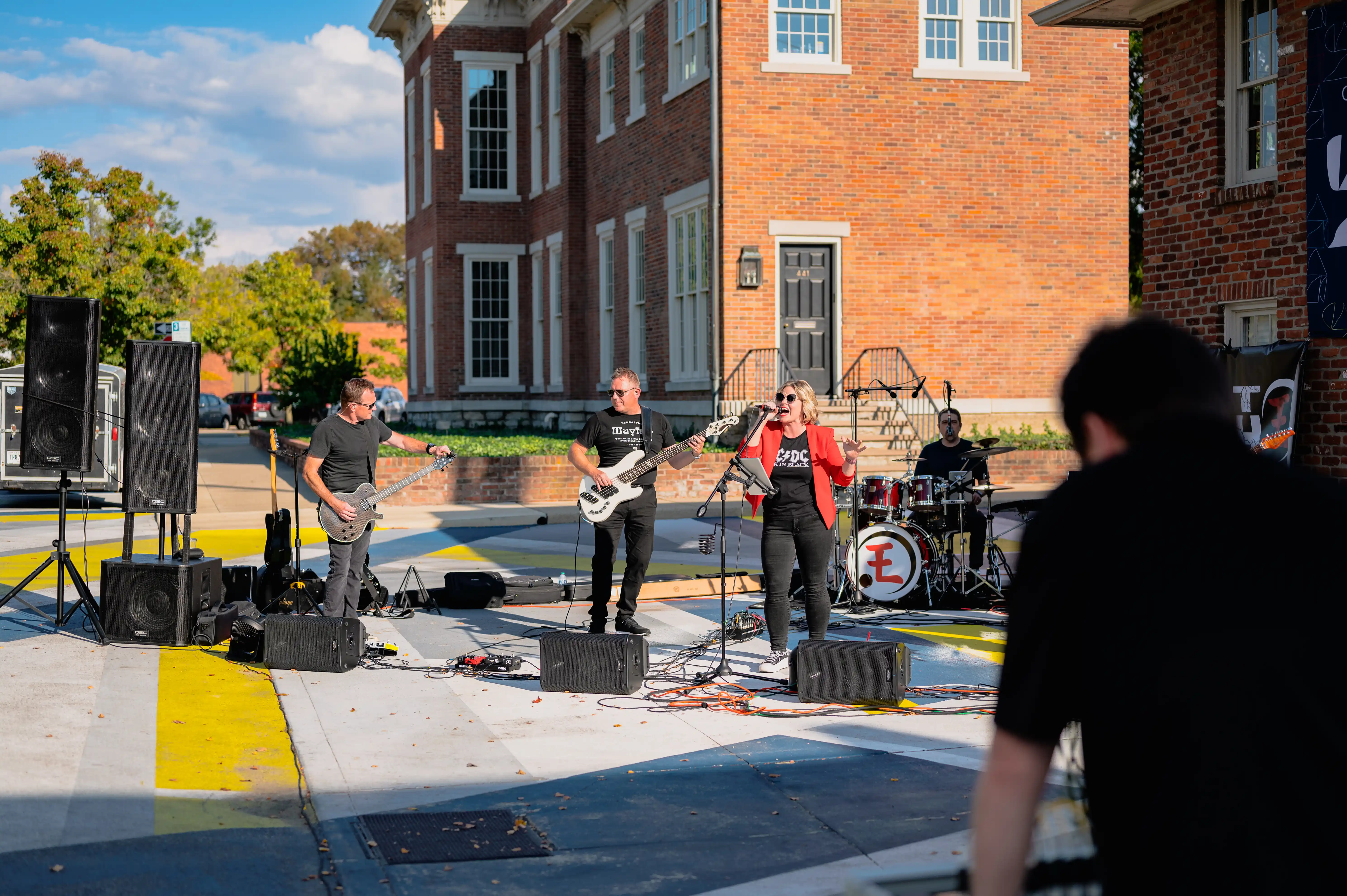 Live band performing outdoors with guitarist, bassist, and drummer in front of a brick building on a sunny day.