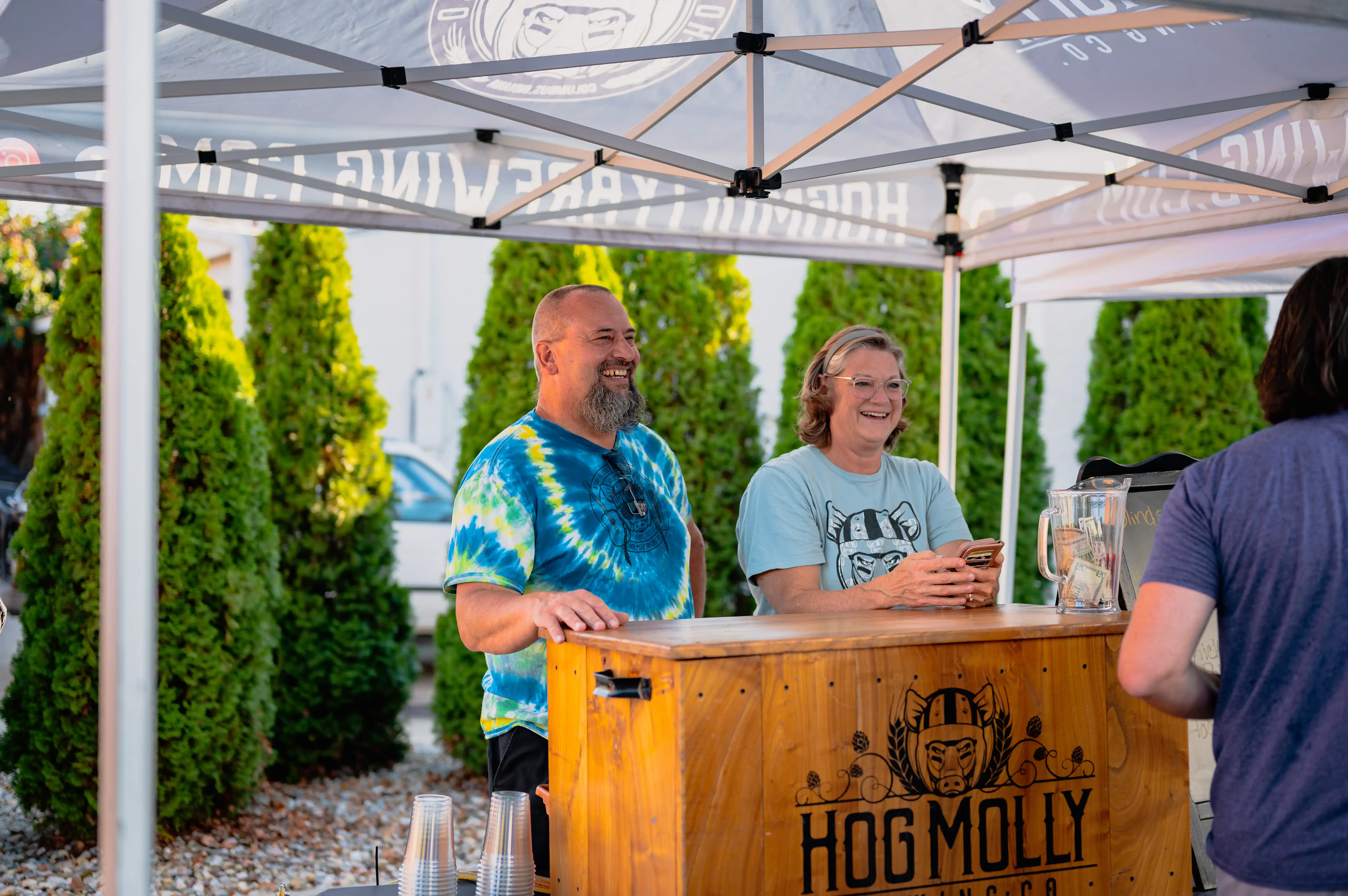 Two people standing under a branded tent, one wearing a tie-dye shirt and the other in gray, smiling and looking towards a customer at their outdoor vendor booth with the sign "HOG MOLLY".