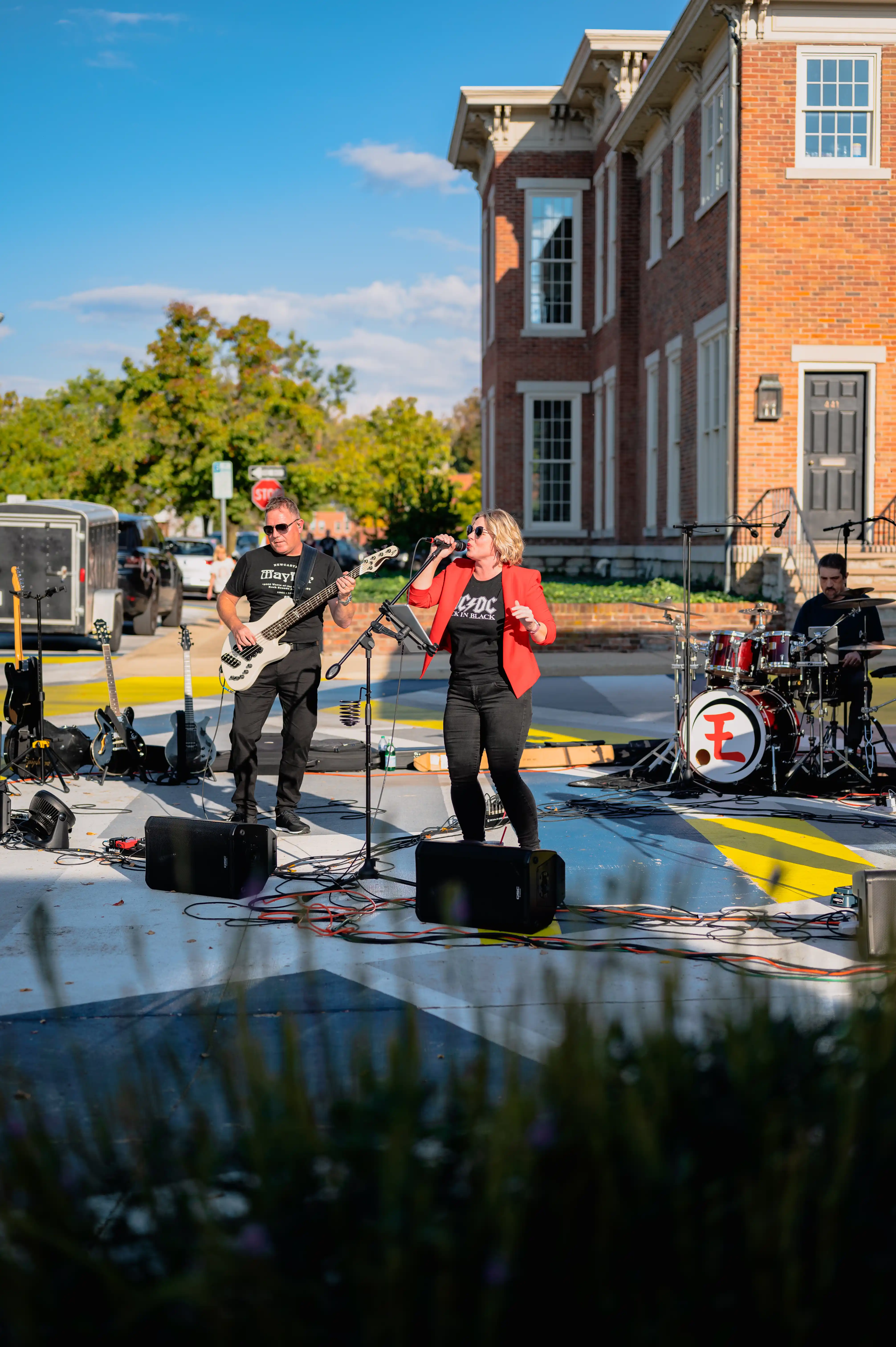 Band performing outdoors on a sunny day with a brick building and trees in the background.