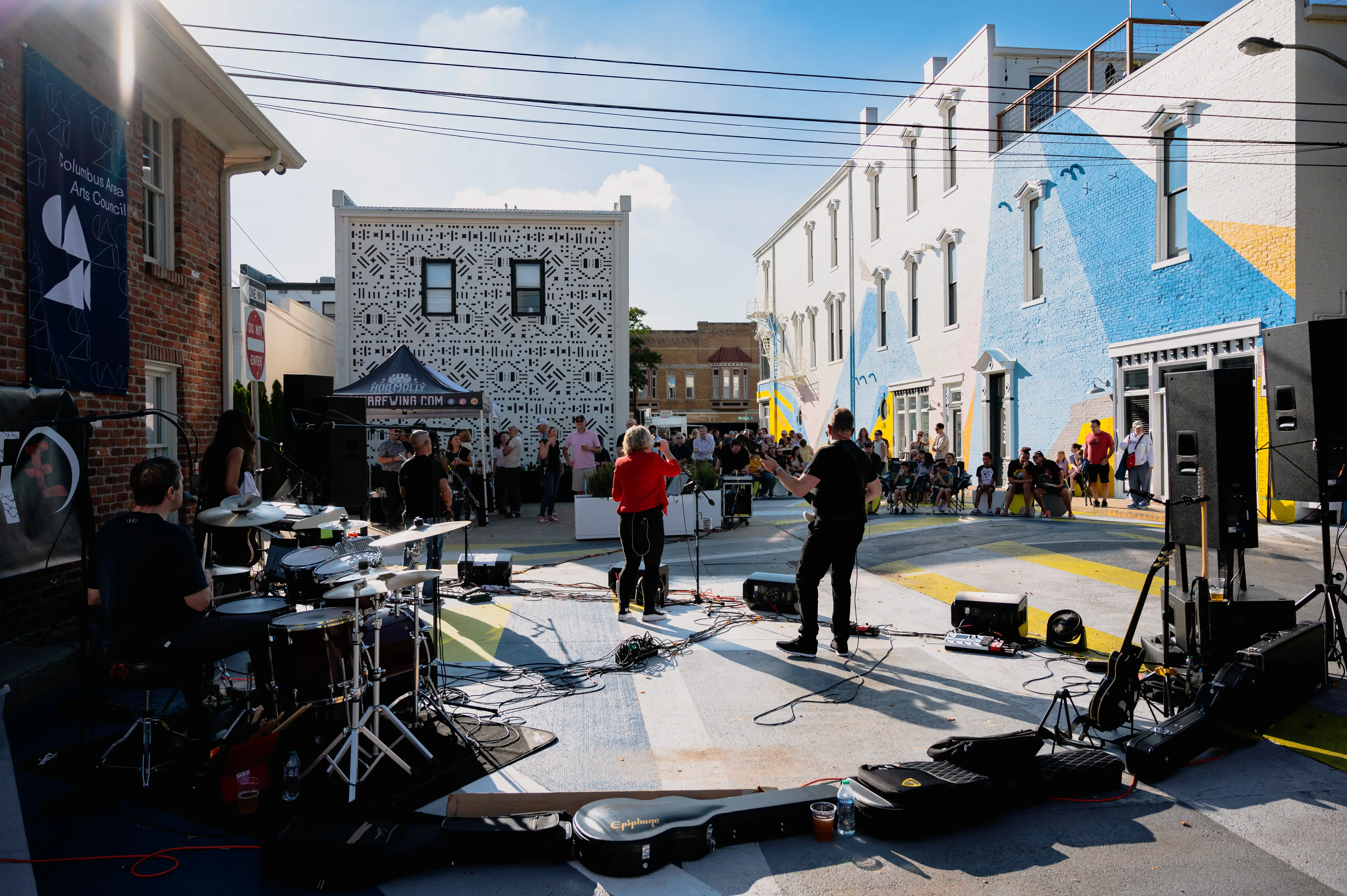 Outdoor live music event with audience watching a band perform on a small street stage, surrounded by buildings with mural art.