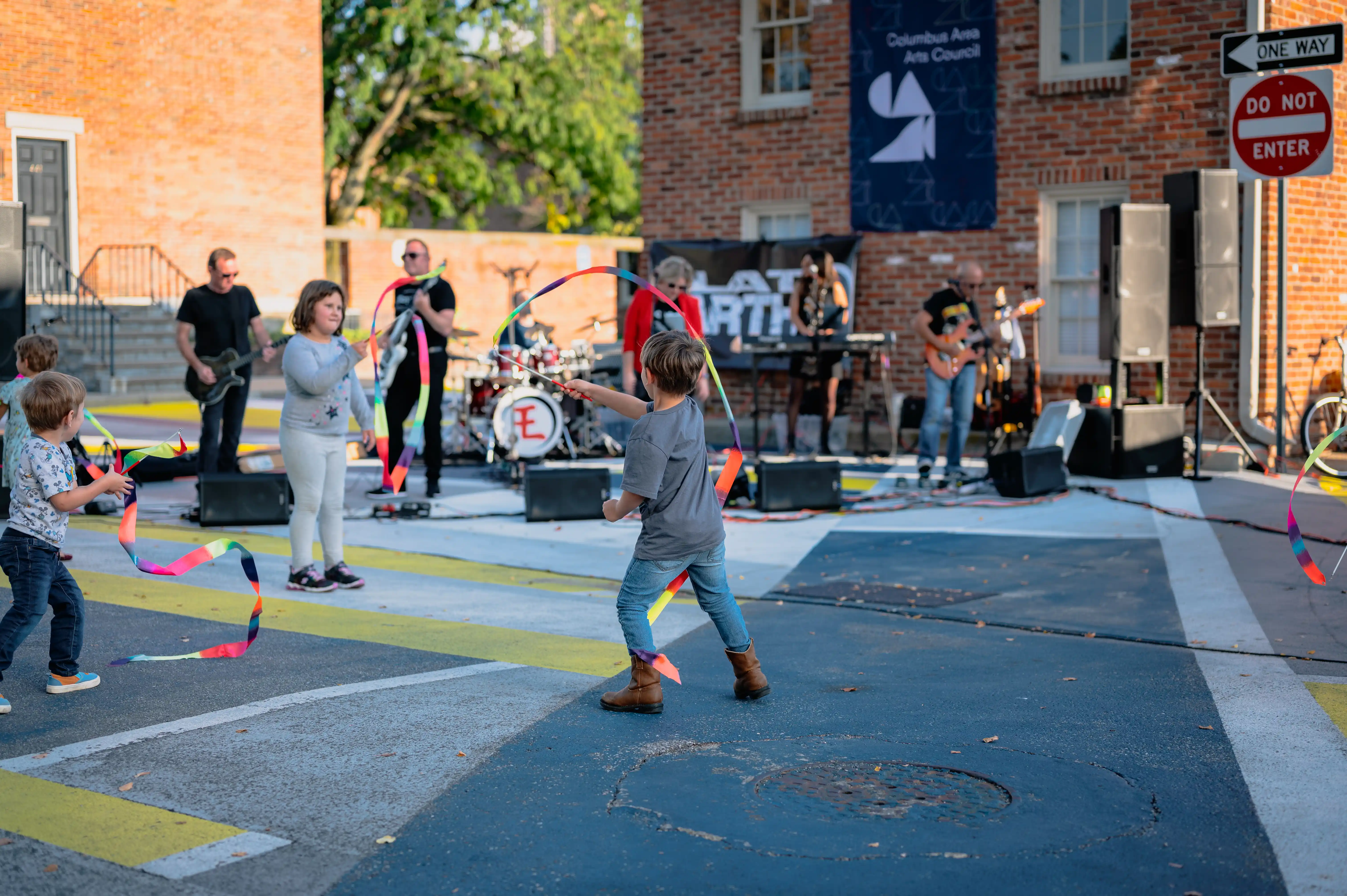 Children playing with hula hoops on a street at an outdoor event with a live band performing in the background.