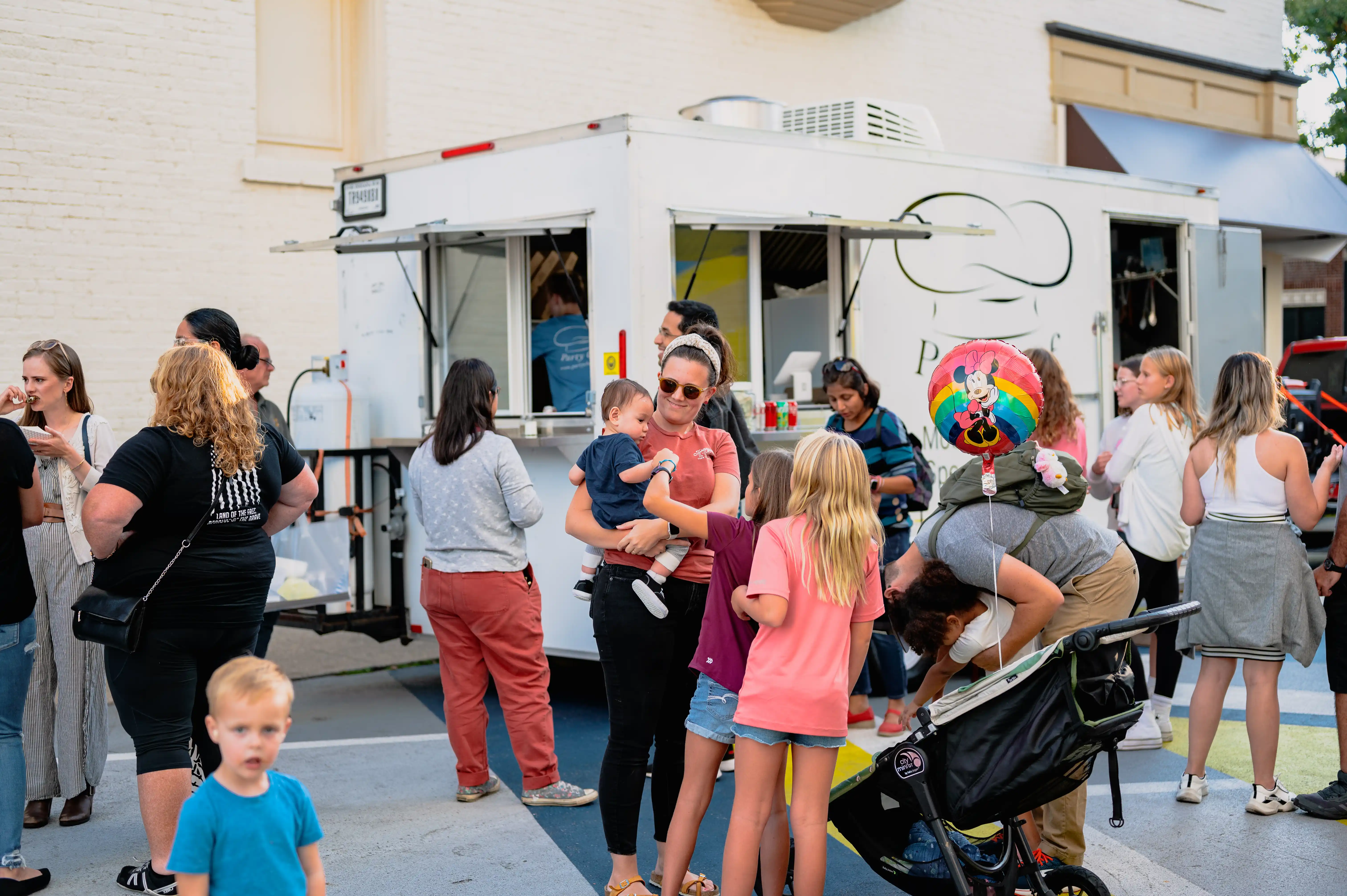 A bustling street food scene with people waiting in line at a food truck, talking and enjoying the atmosphere, some with children and strollers.
