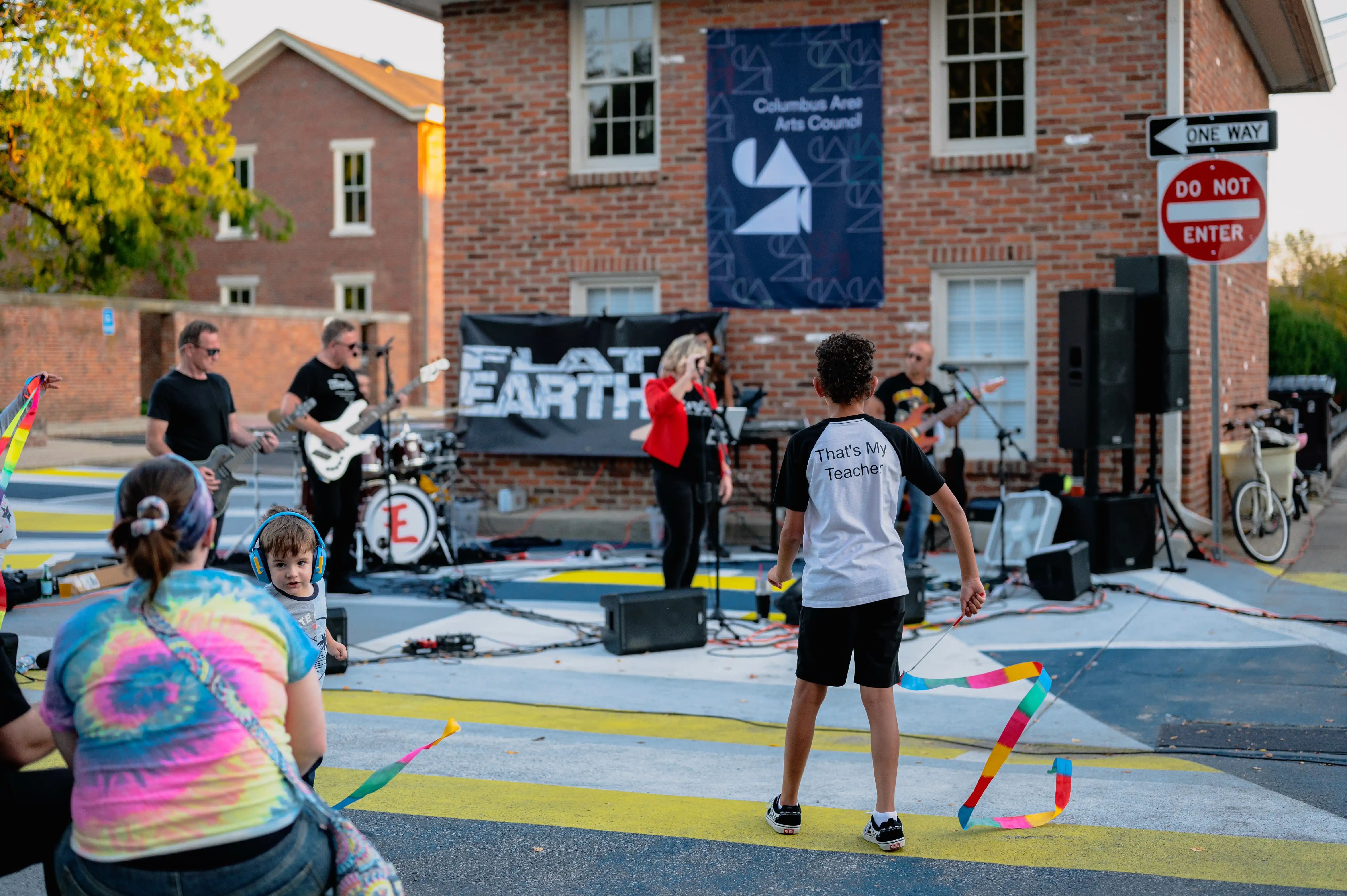 Band performing on an outdoor stage with a small audience, featuring a guitarist, vocalist, and drummer, and a person in a tie-dye shirt watching.