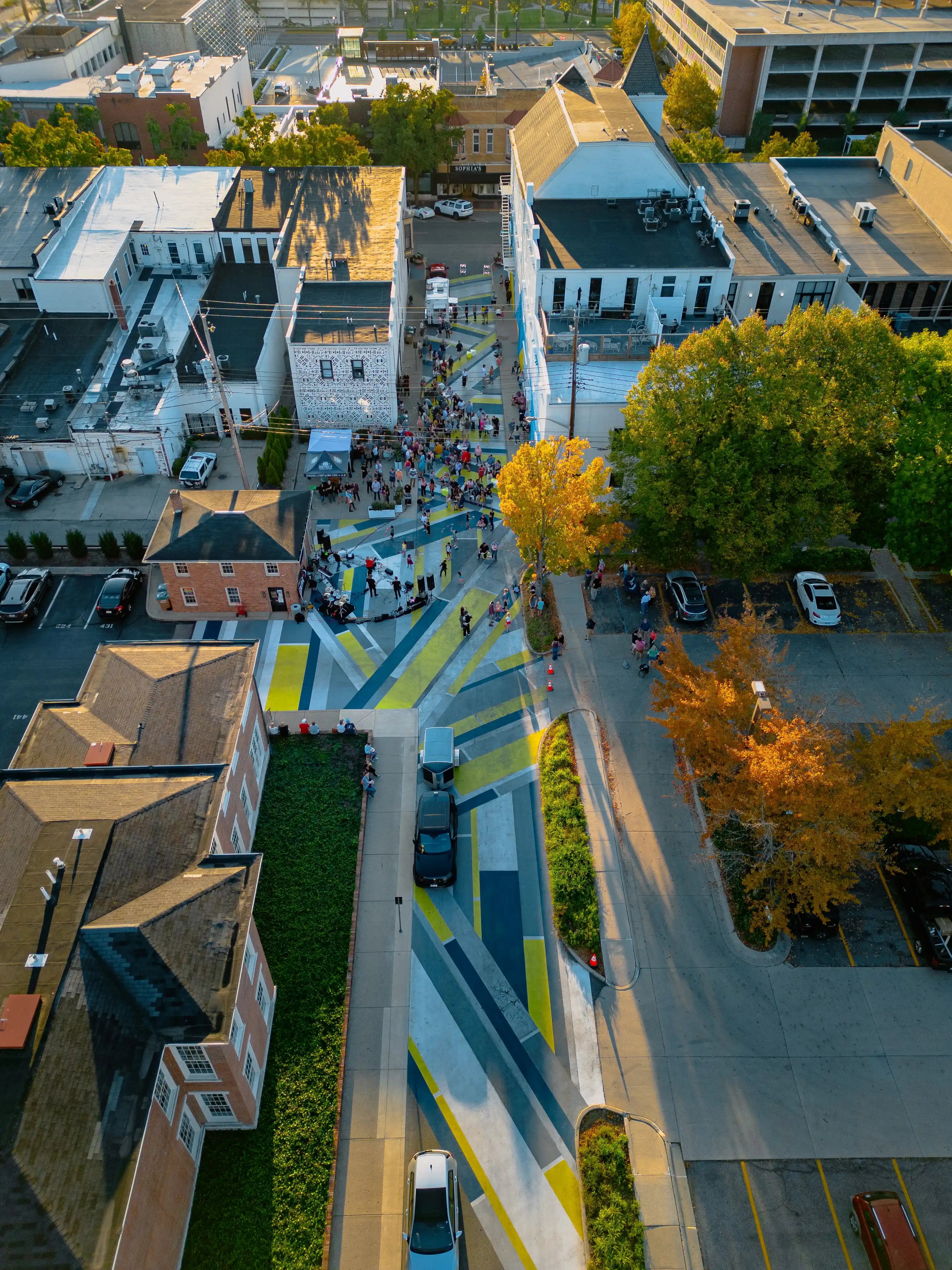 Aerial view of a busy city street intersection with cars, pedestrians, and autumn-colored trees during early evening.