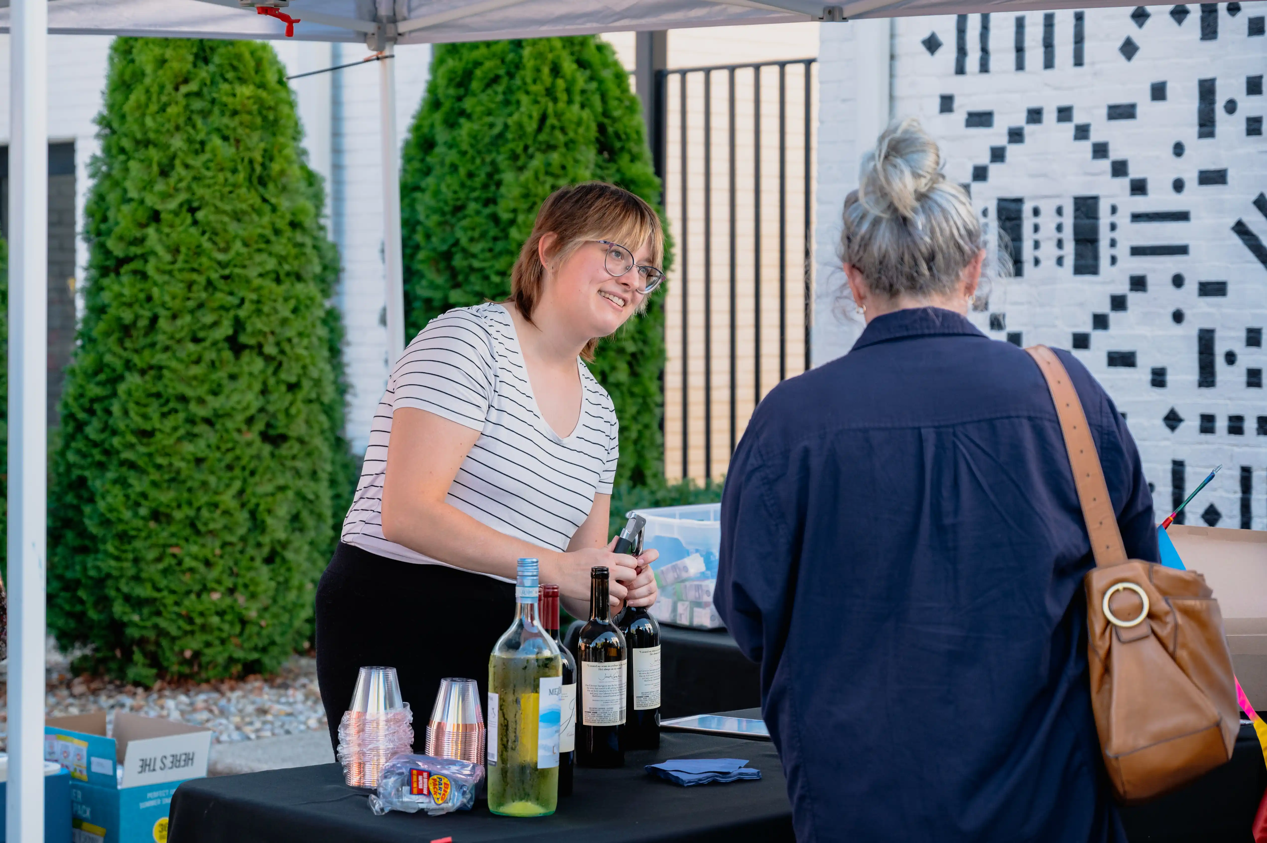 A woman smiling while serving wine at an outdoor tasting booth with another person standing in front.