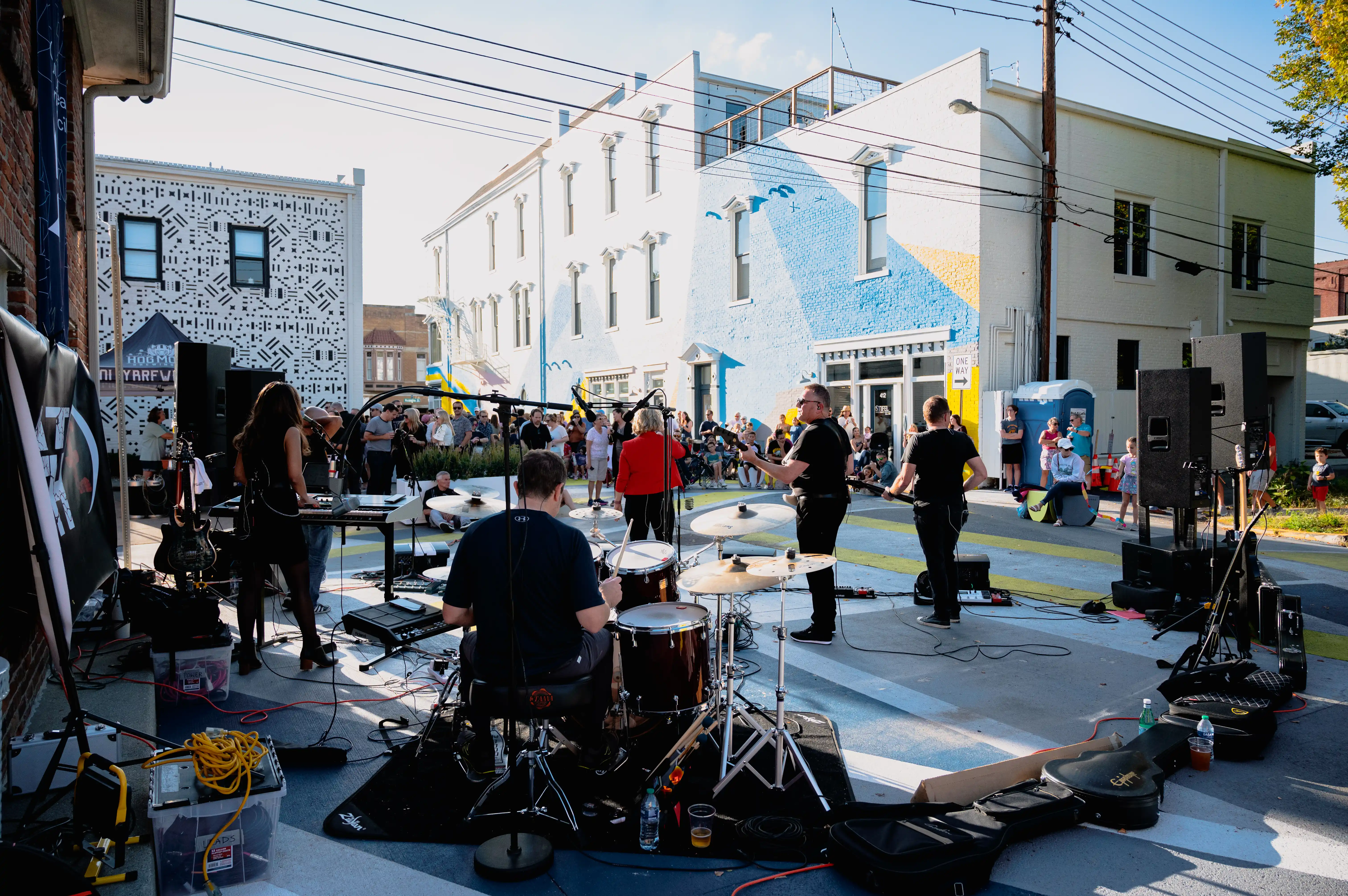 A live band performs on a street stage in front of an audience during a sunny outdoor event, with buildings and urban street art in the background.