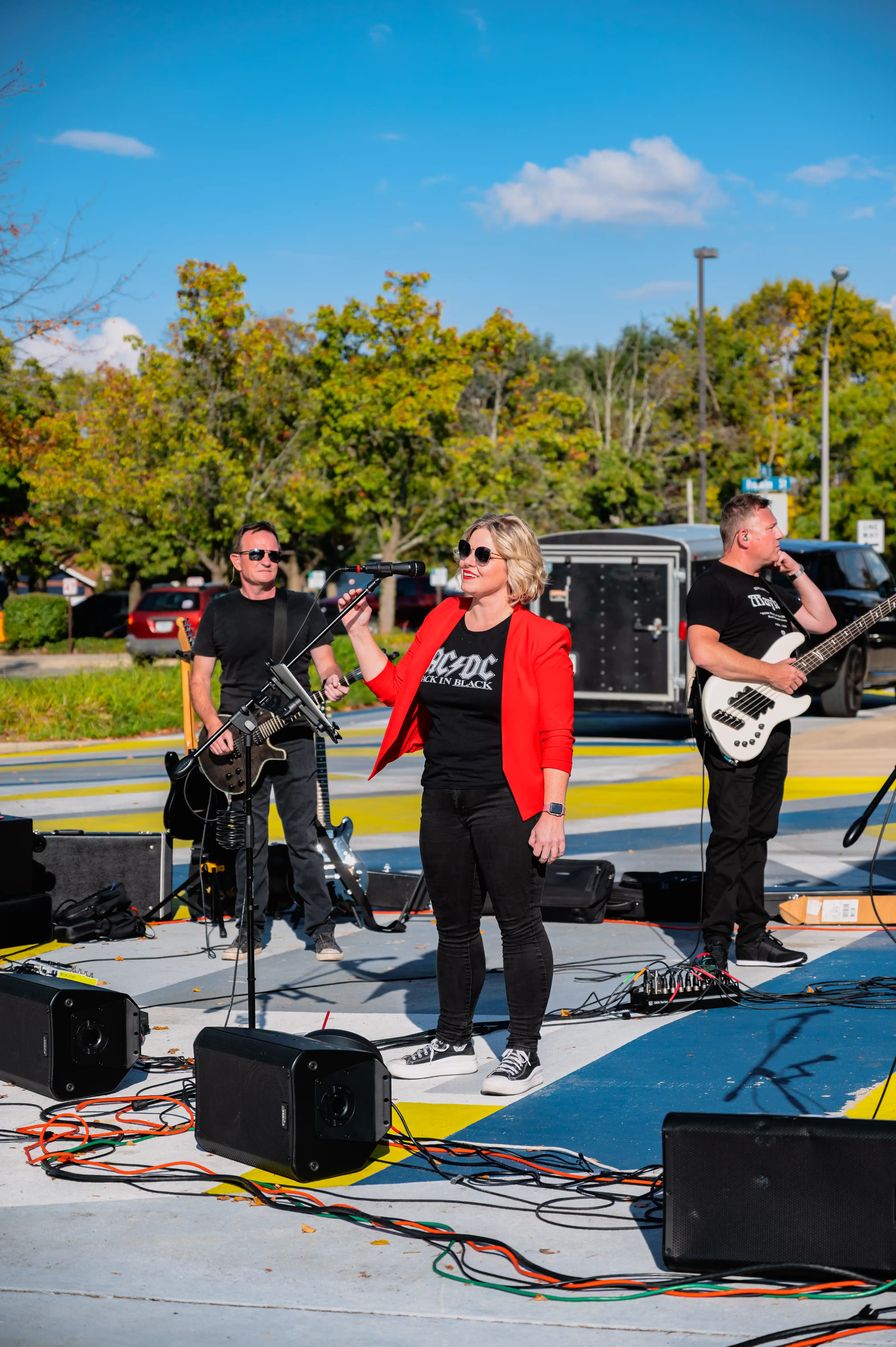 Band performing on an outdoor stage in daytime, with a female singer in front and male guitarists and bassist in the background, with scenic autumn trees and blue sky.