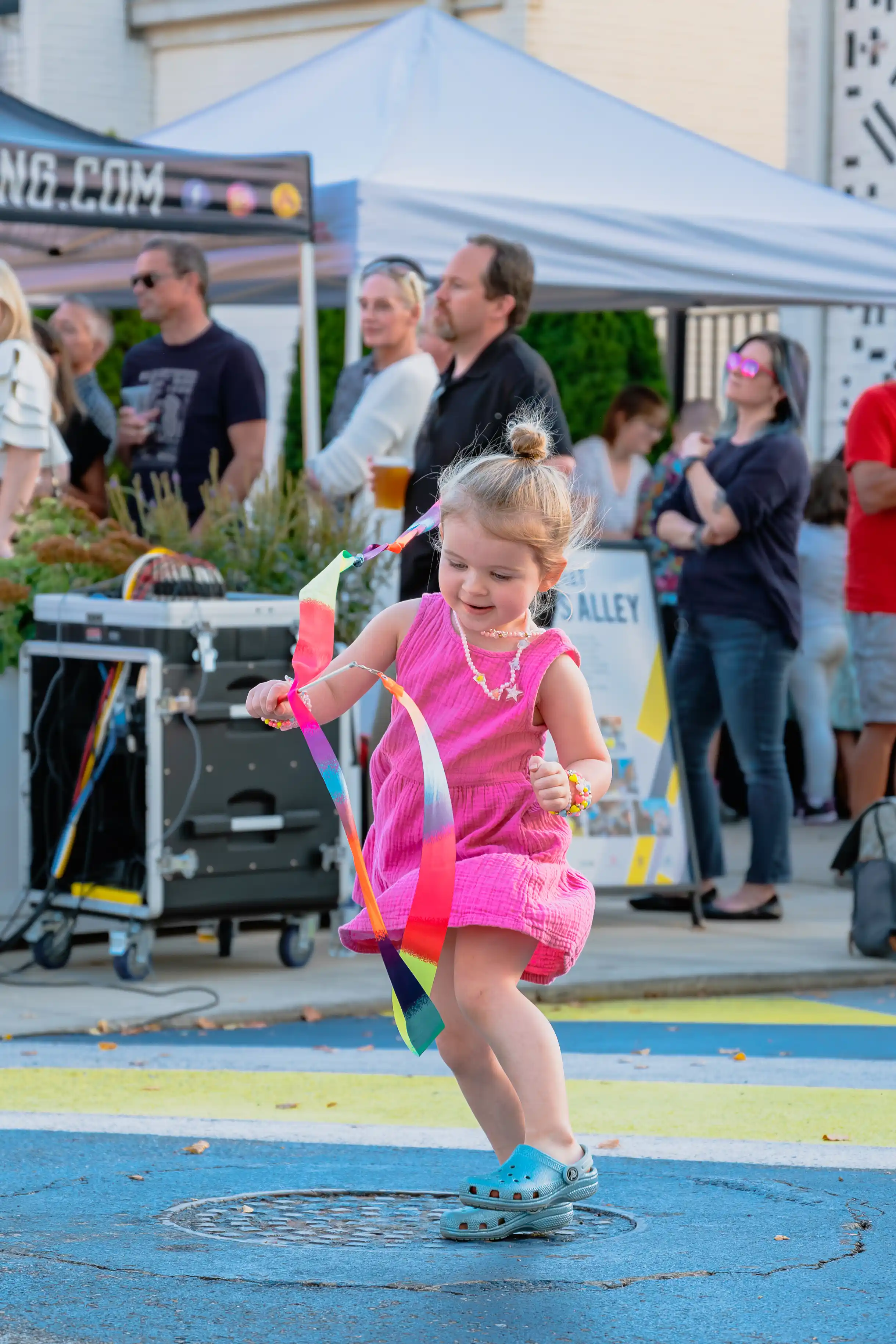 A young girl in a pink dress playfully dancing with colorful ribbons at a street festival with onlookers in the background.