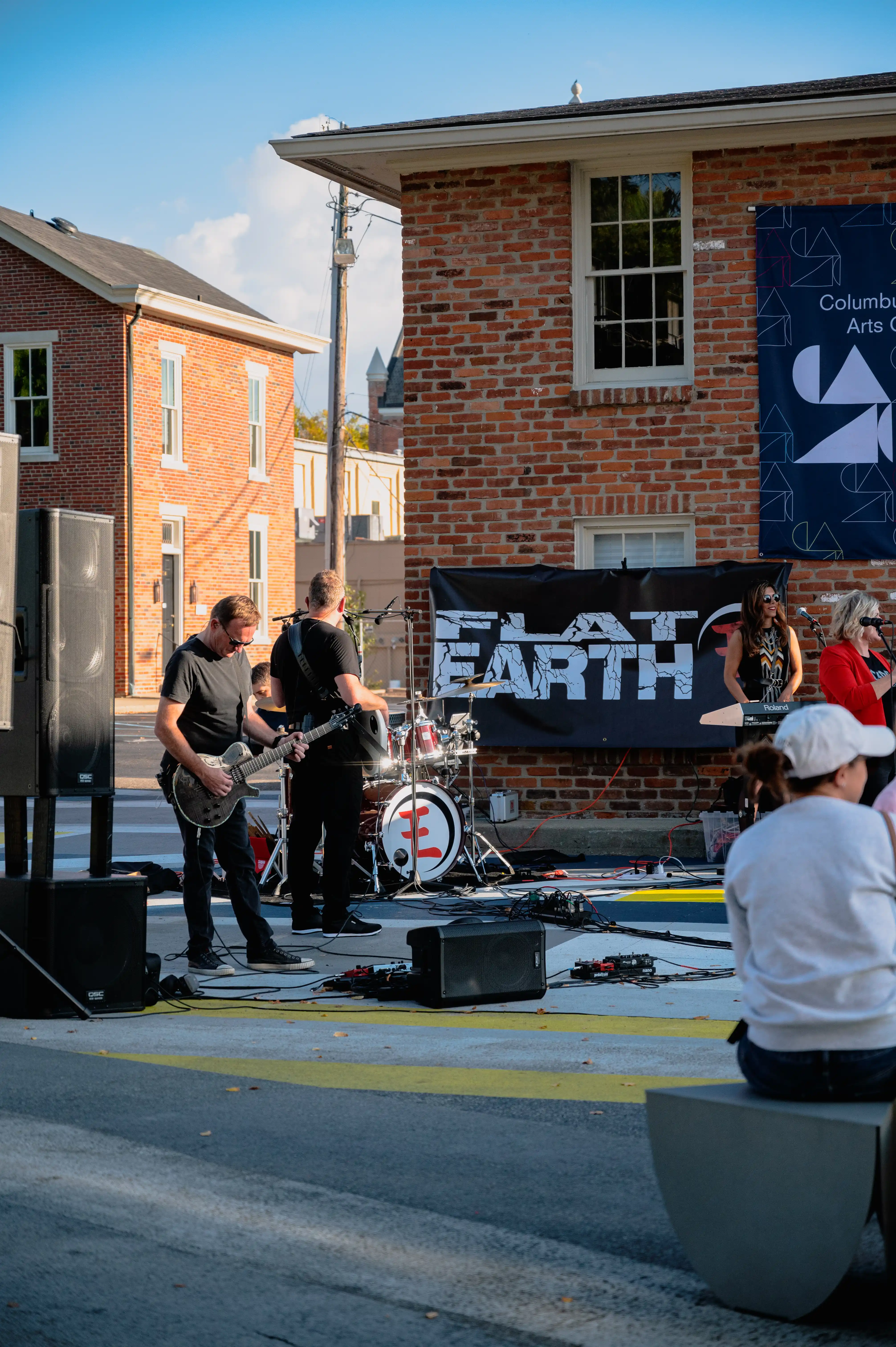 A local band performing at an outdoor event with a banner reading "FLAT EARTH" in the background, spectators in the foreground, and brick buildings under a clear sky.