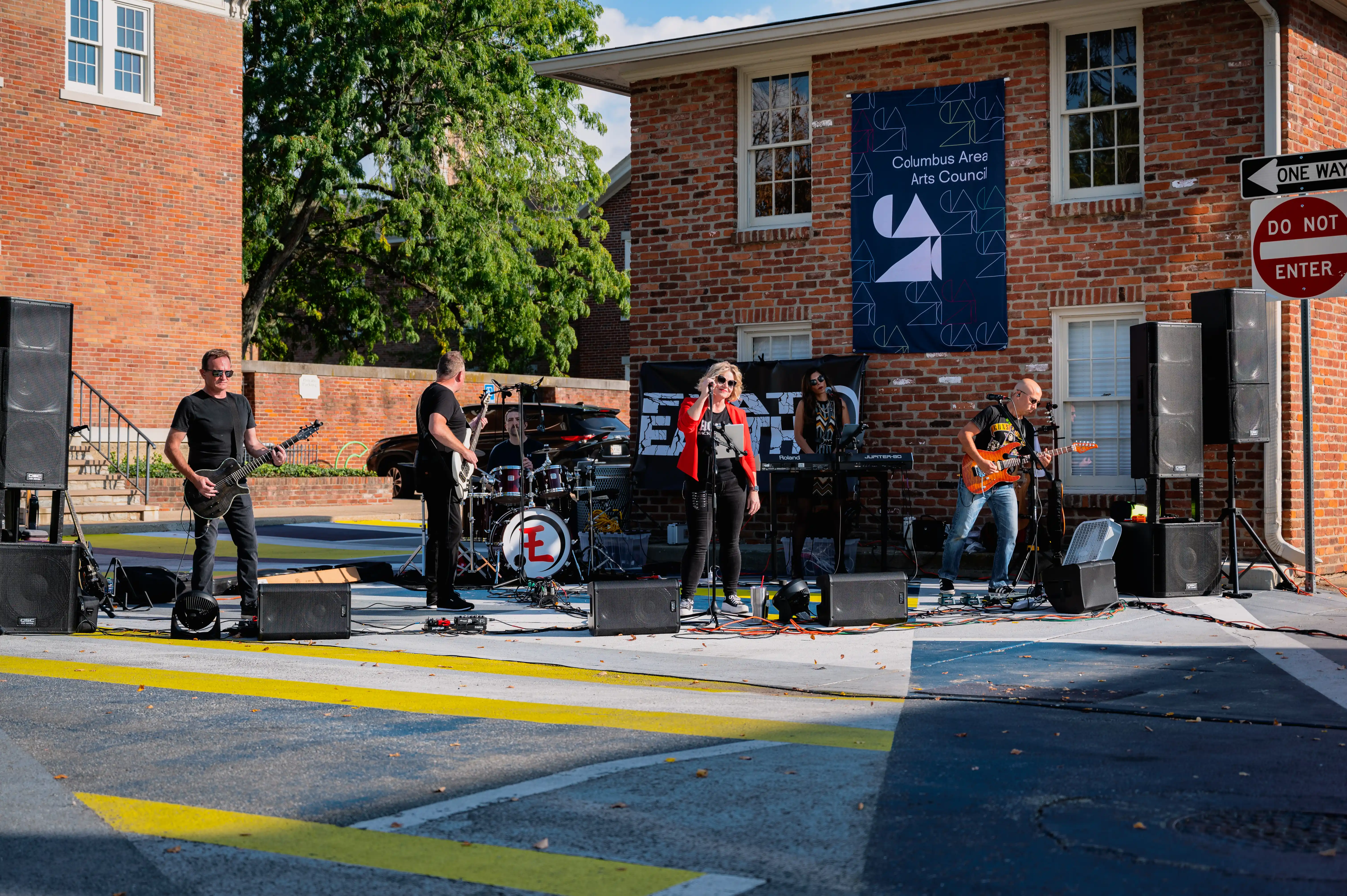 A live band performing outdoors with a singer, guitarists, a drummer, and keyboard player in front of a Columbus Area Arts Council sign.