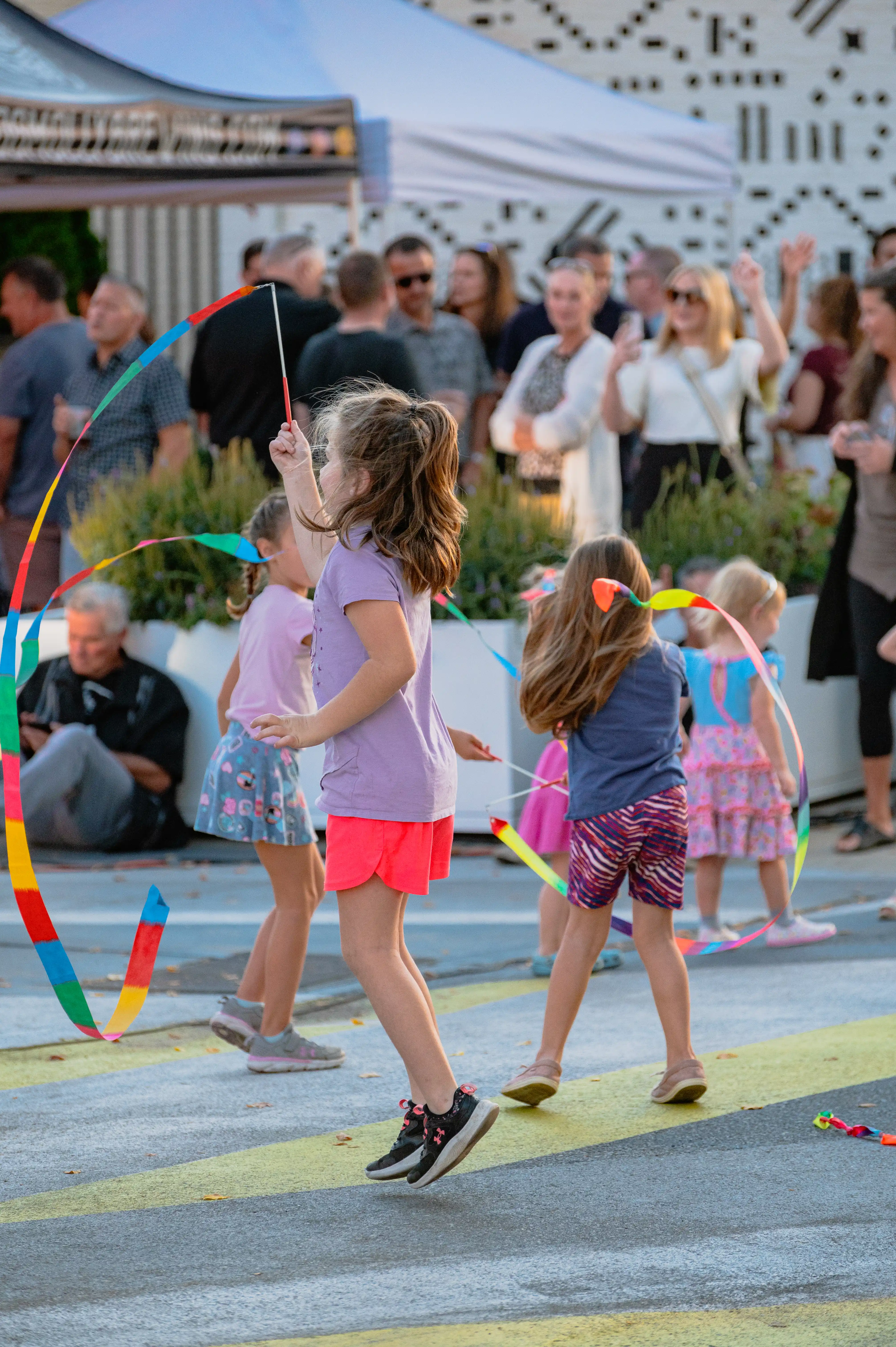 Children playing with colorful ribbons at an outdoor event with onlookers in the background.