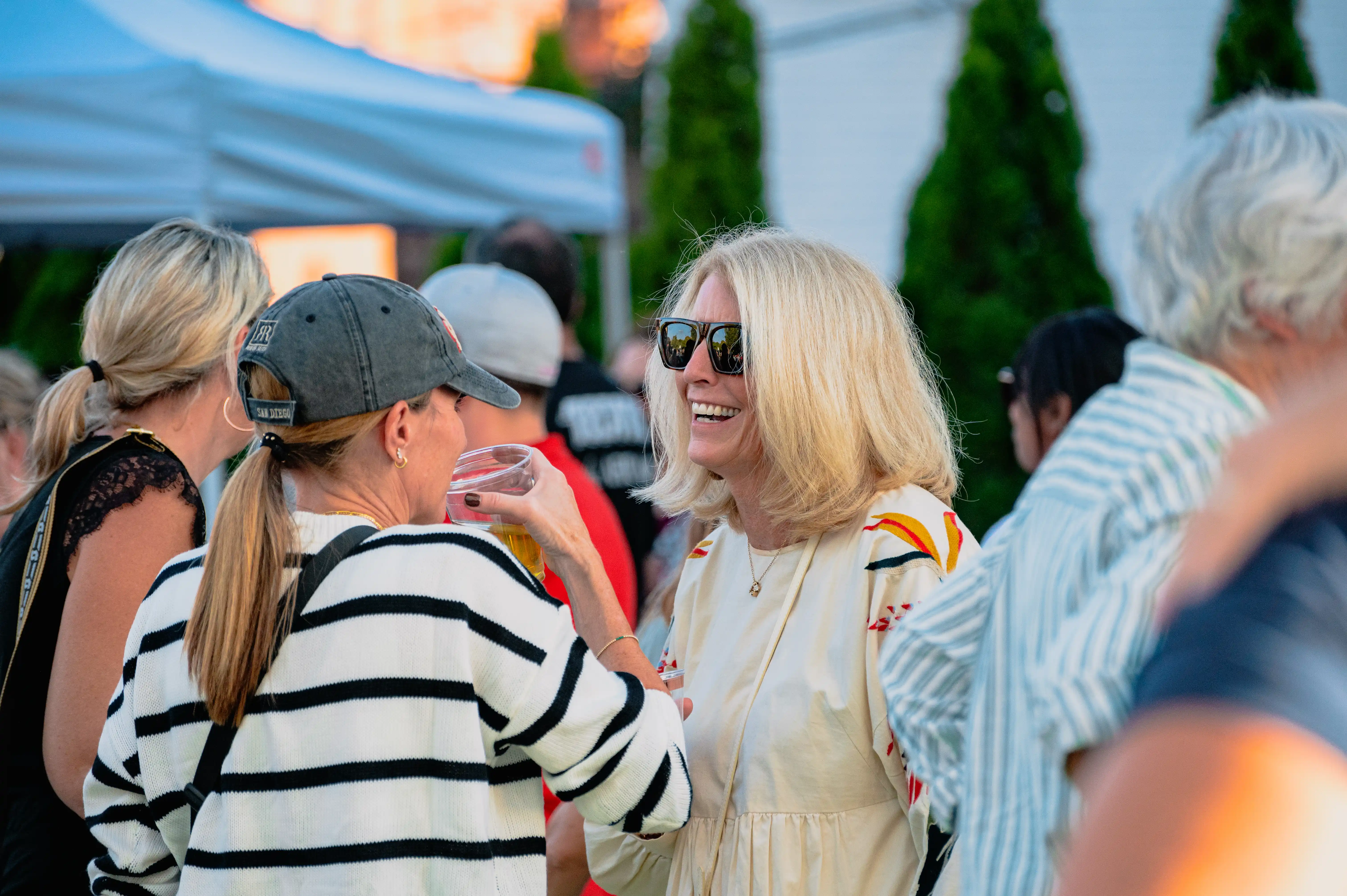 People socializing at an outdoor event with a woman smiling and conversing in the foreground.
