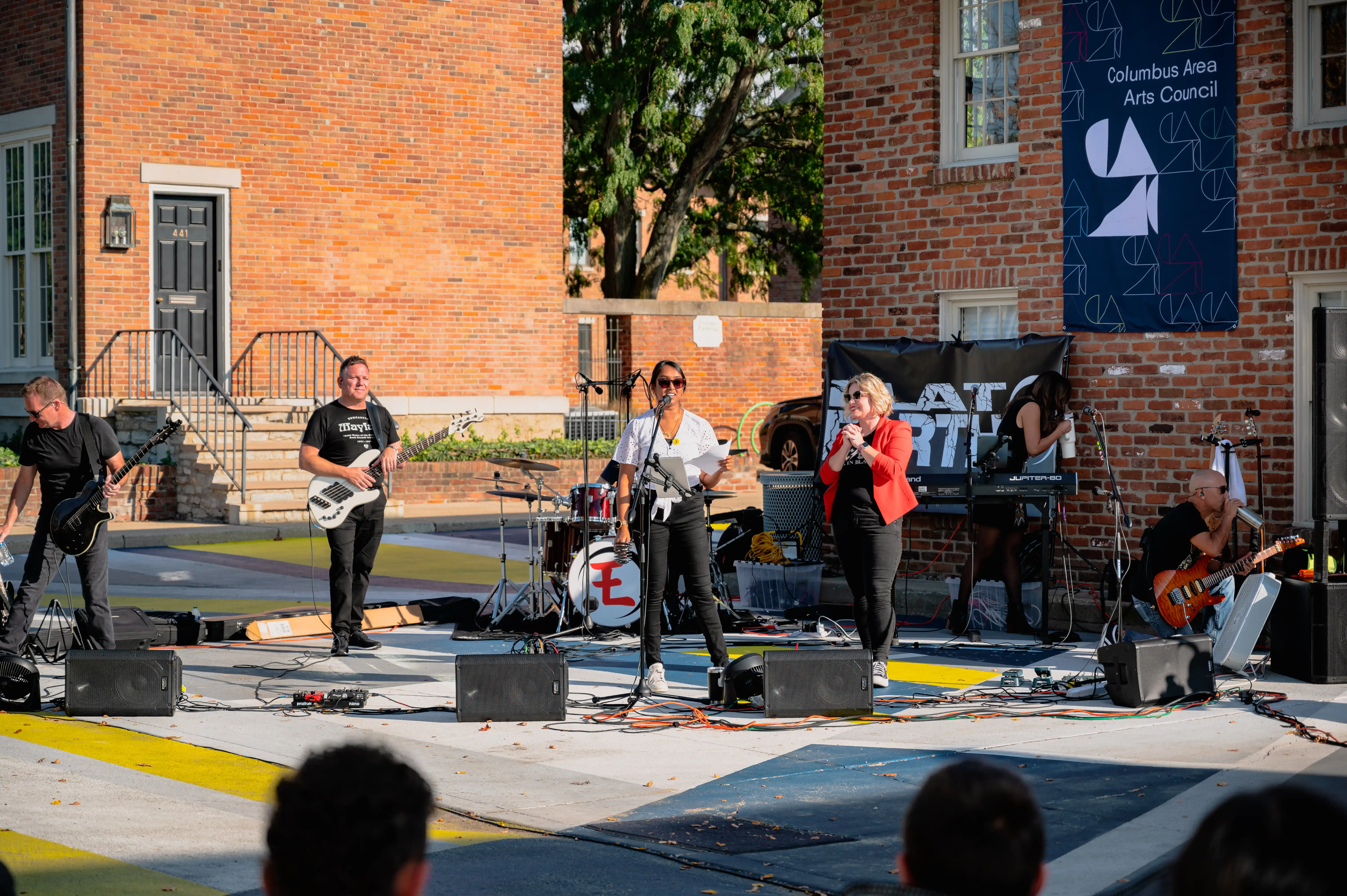Band performing on an outdoor stage in front of a brick building with a banner that reads "Columbus Area Arts Council"