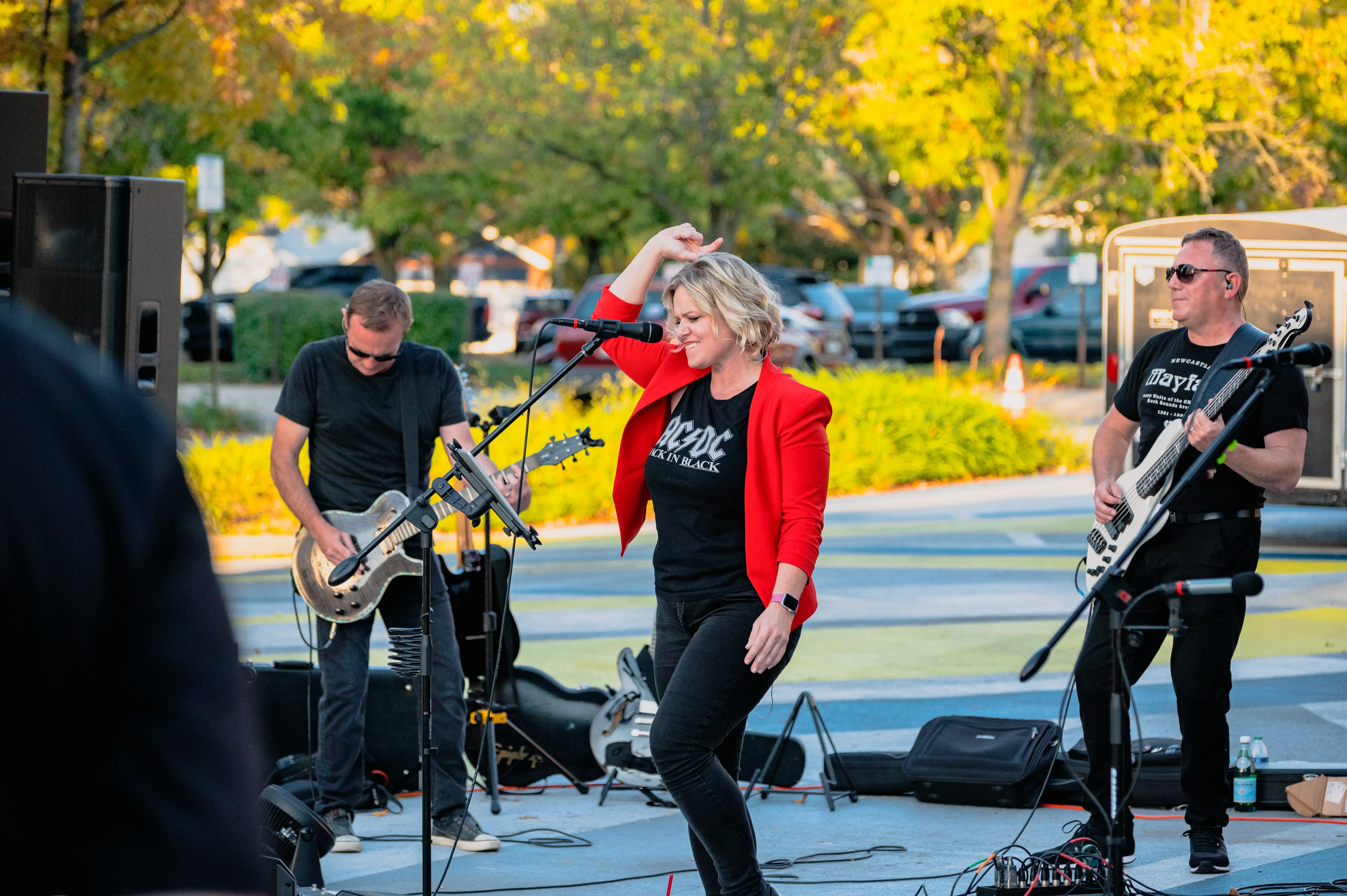 A band performing outdoors with a female singer in red jacket and two guitarists.