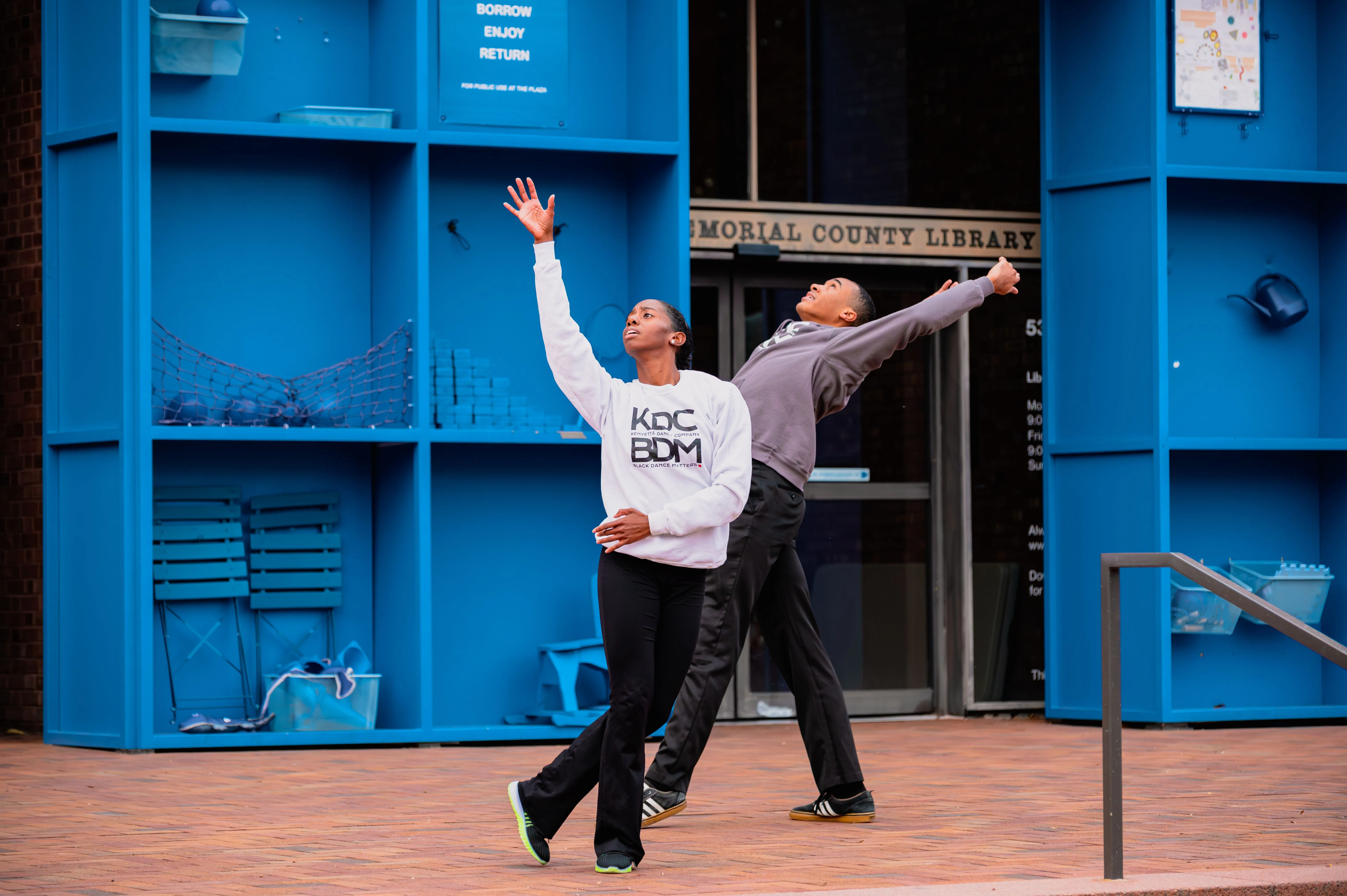 Two individuals in casual attire joyfully dancing outside a blue building with "Public Library" signage.