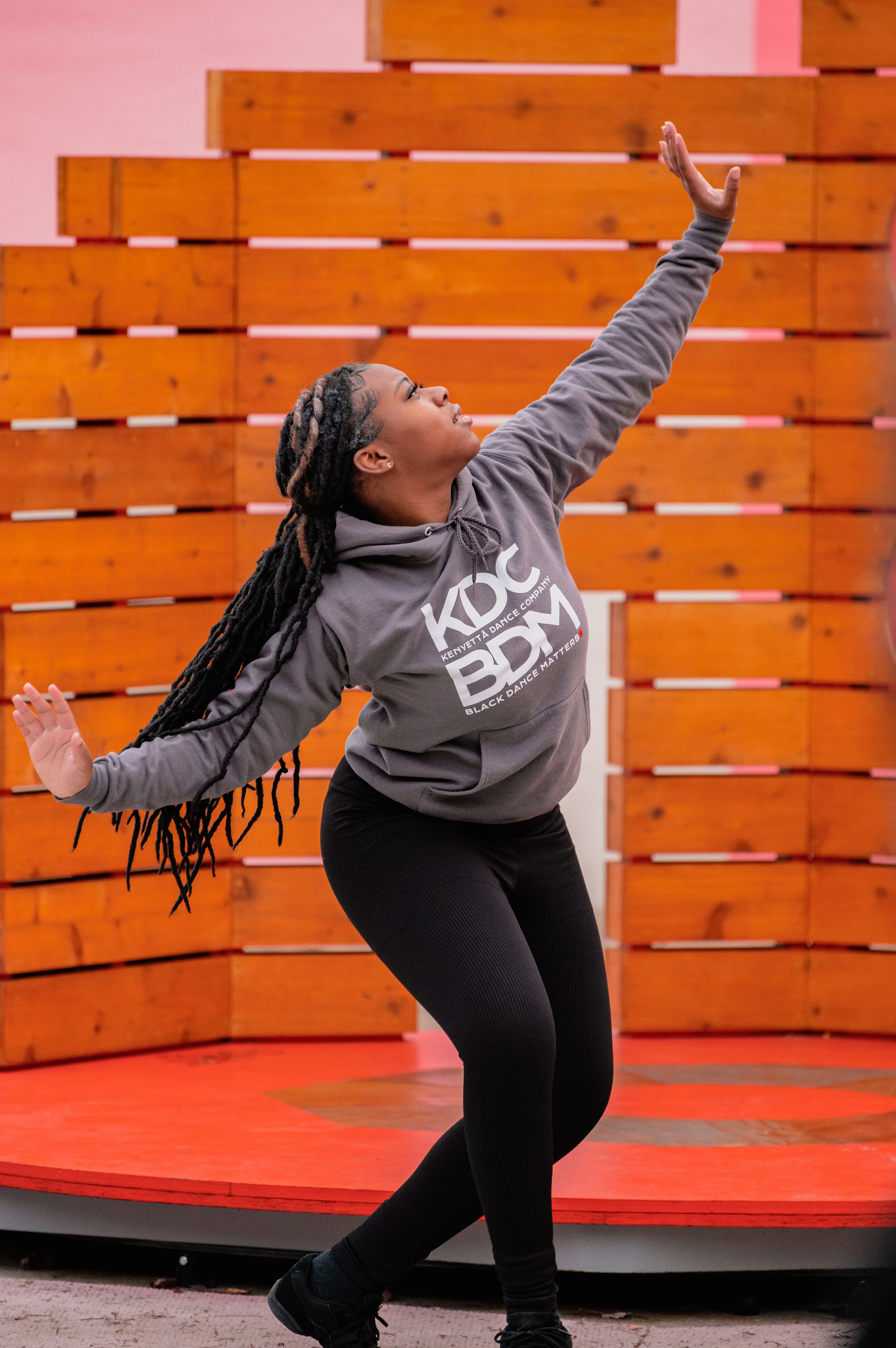 Woman with braids wearing casual clothes dancing and reaching upwards in front of an orange pallet wall.
