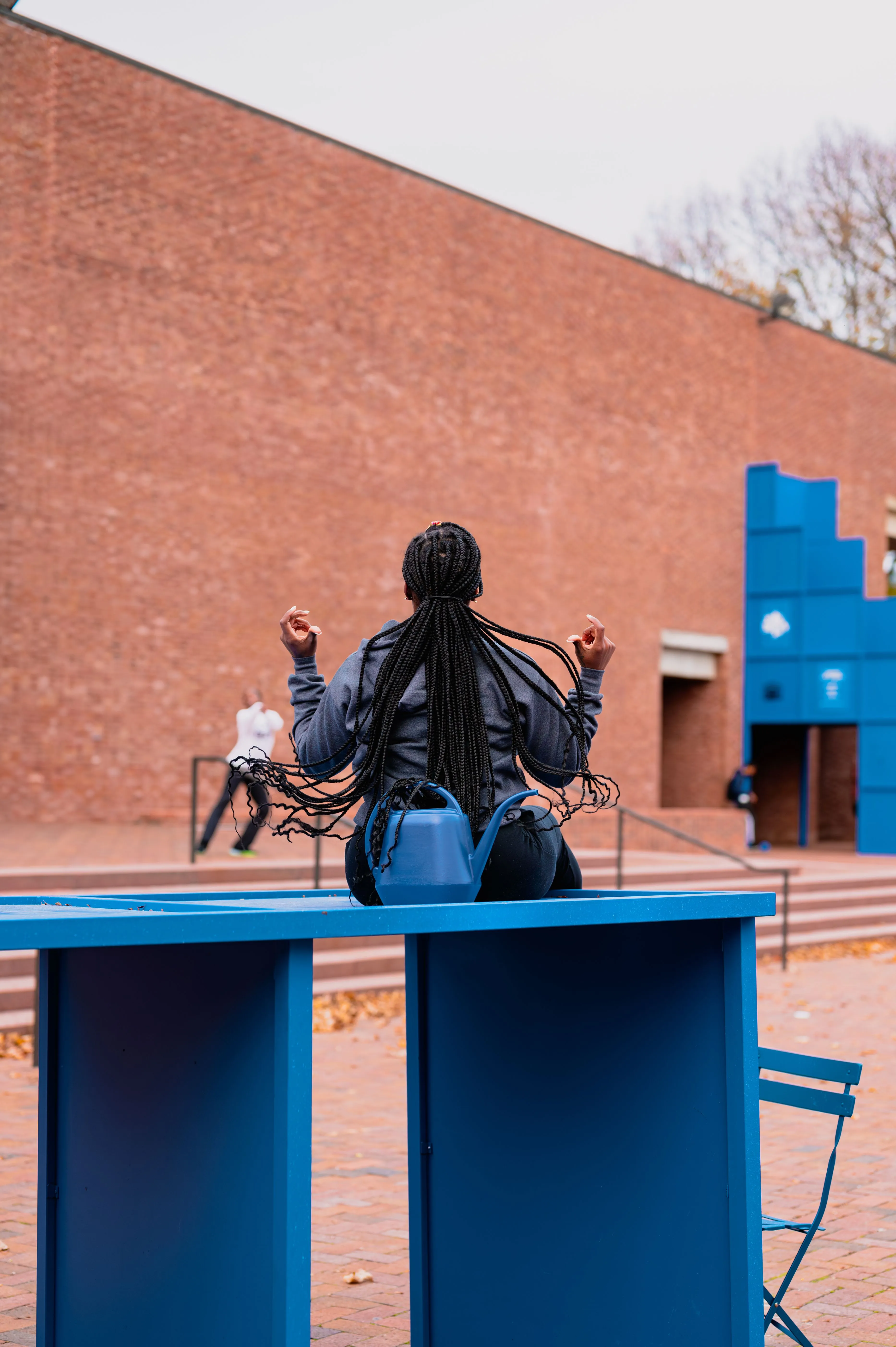 Person sitting on a blue ledge in an outdoor urban setting with another individual in the background.