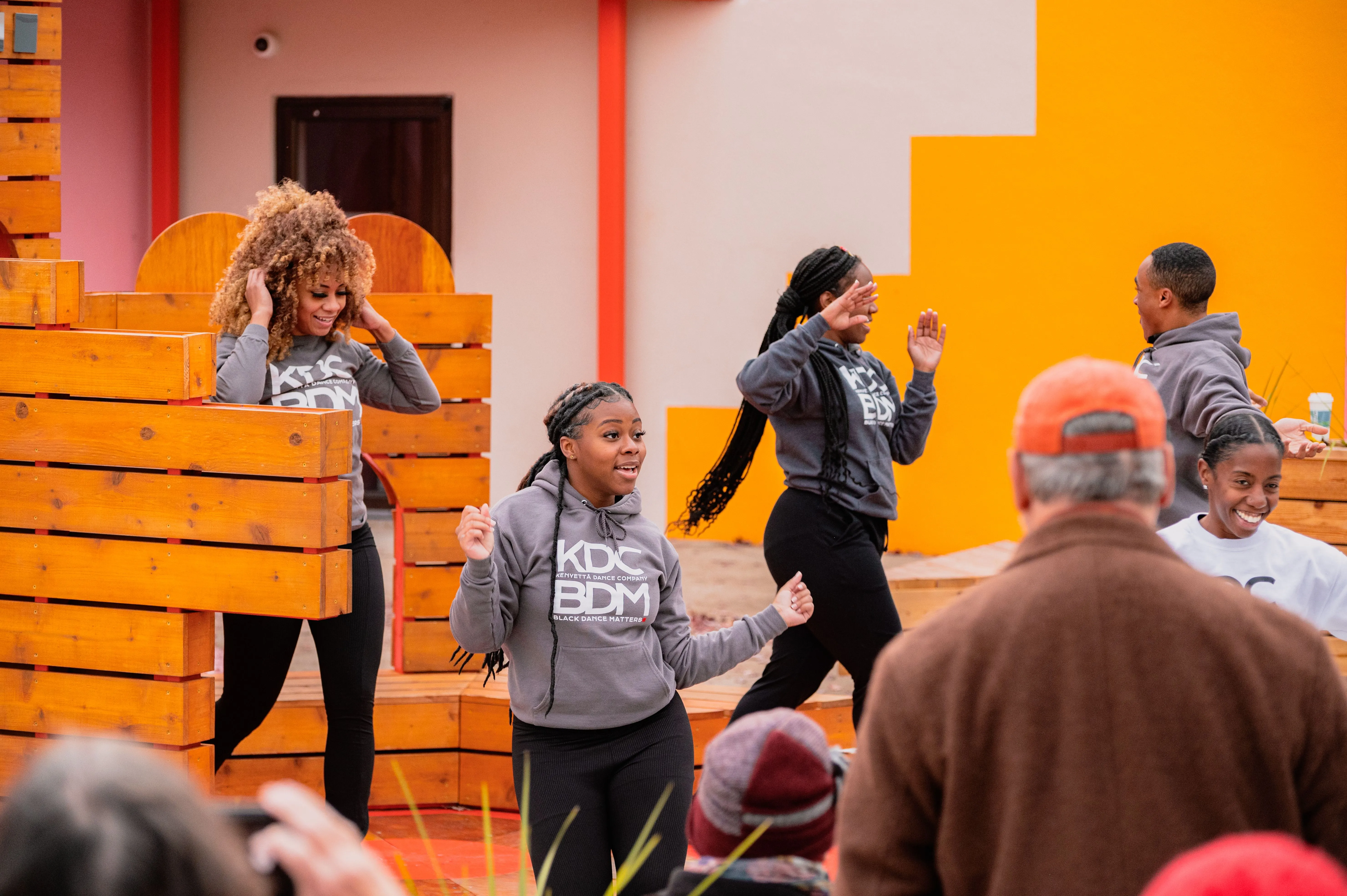 People dancing on the street with a vibrant orange backdrop and wooden pallets.