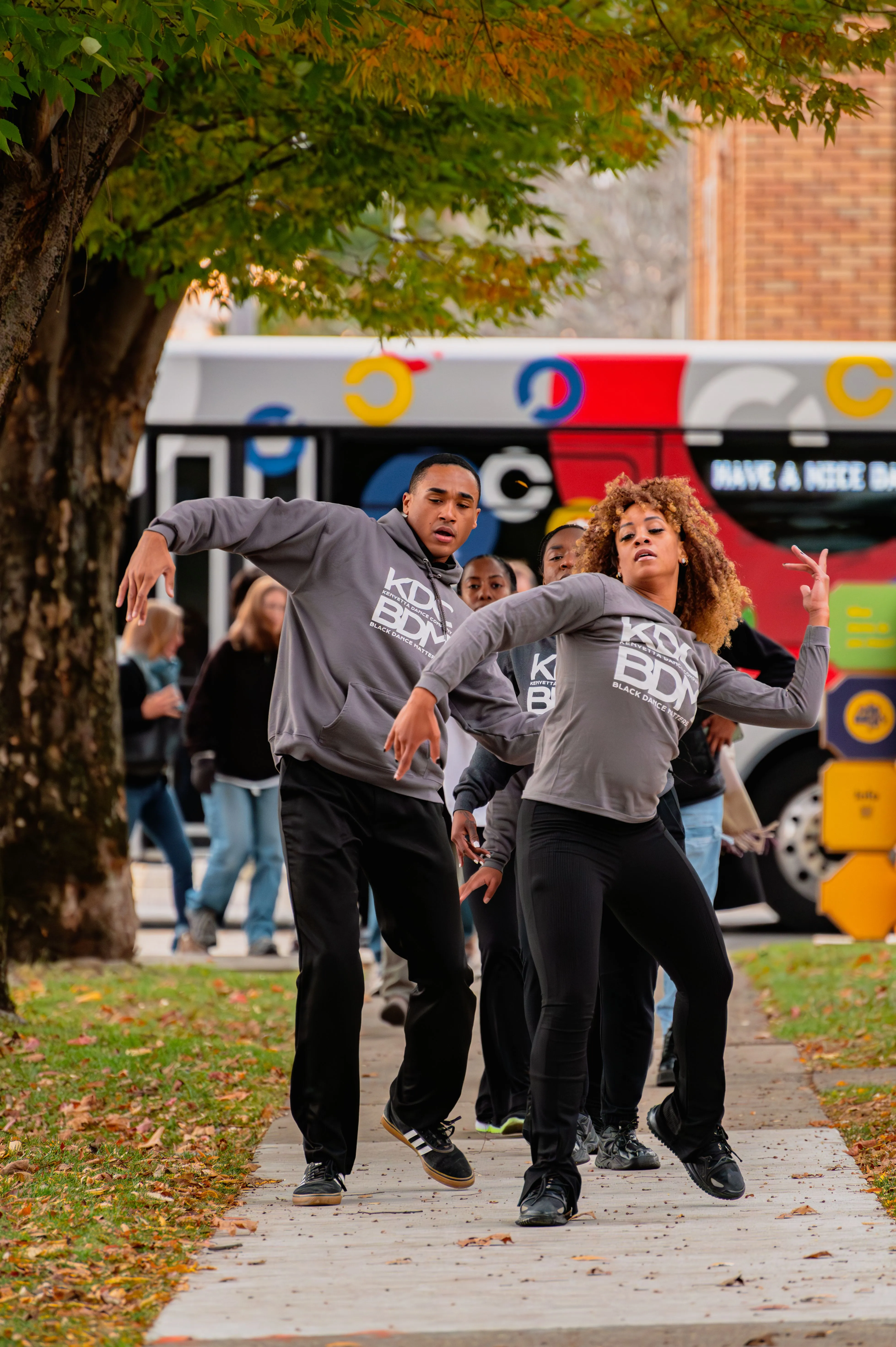 Group of people playfully dancing on a sidewalk with autumn leaves on the ground and a colorful bus in the background.
