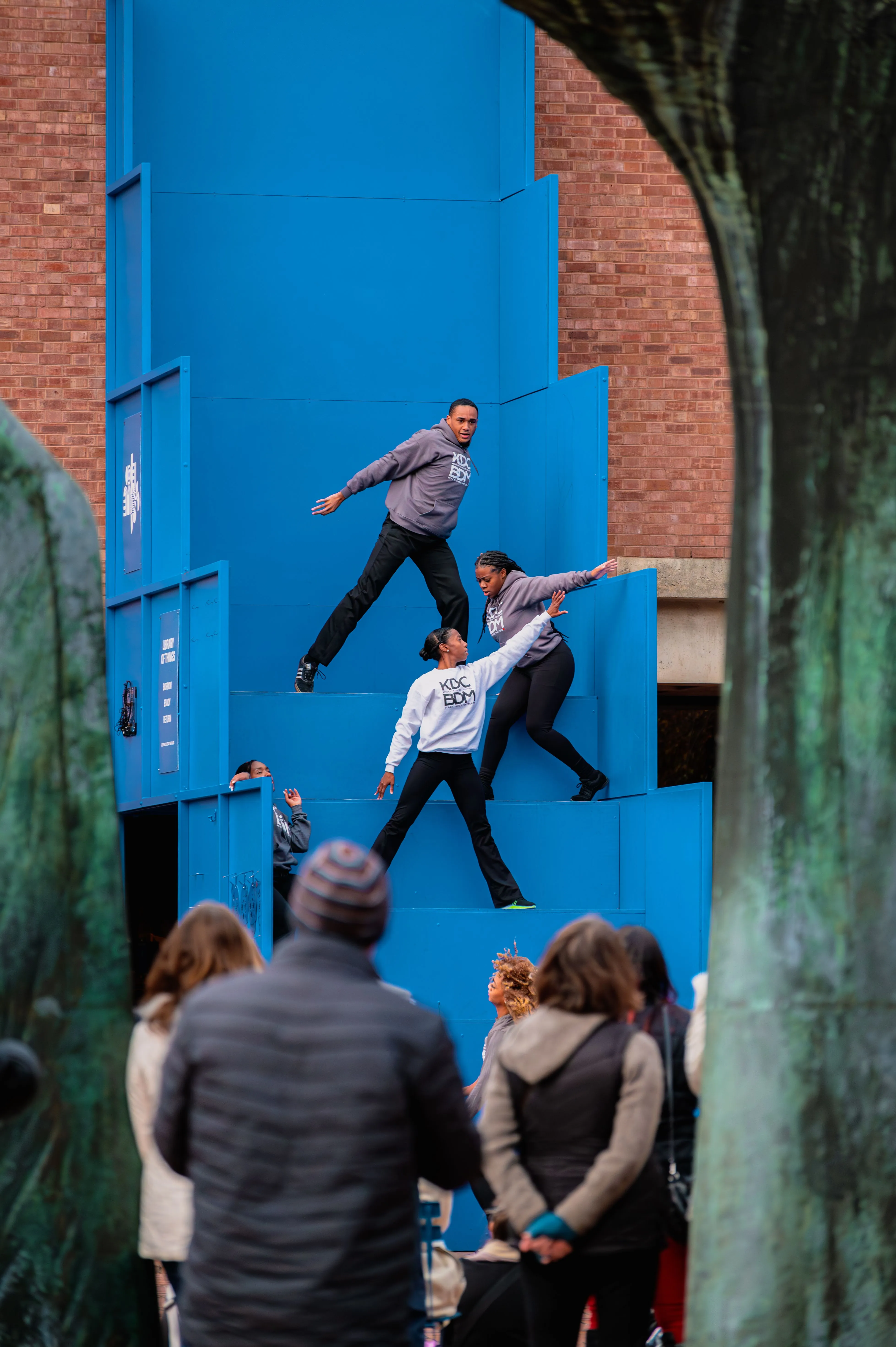 Spectators watching an illusion of two people climbing and defying gravity on a vertical, blue painted street wall.