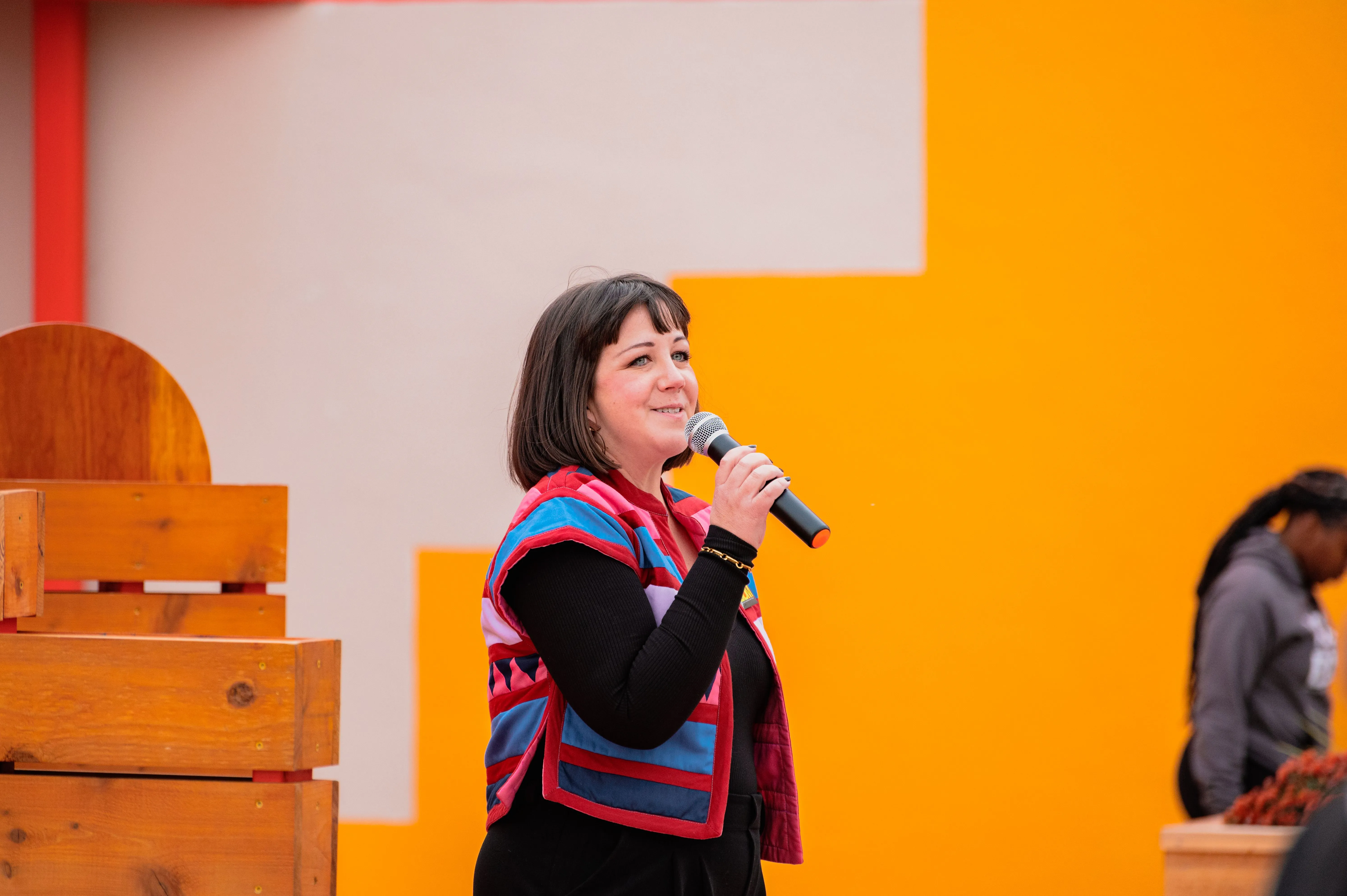 Woman speaking into a microphone at an event with an orange background.