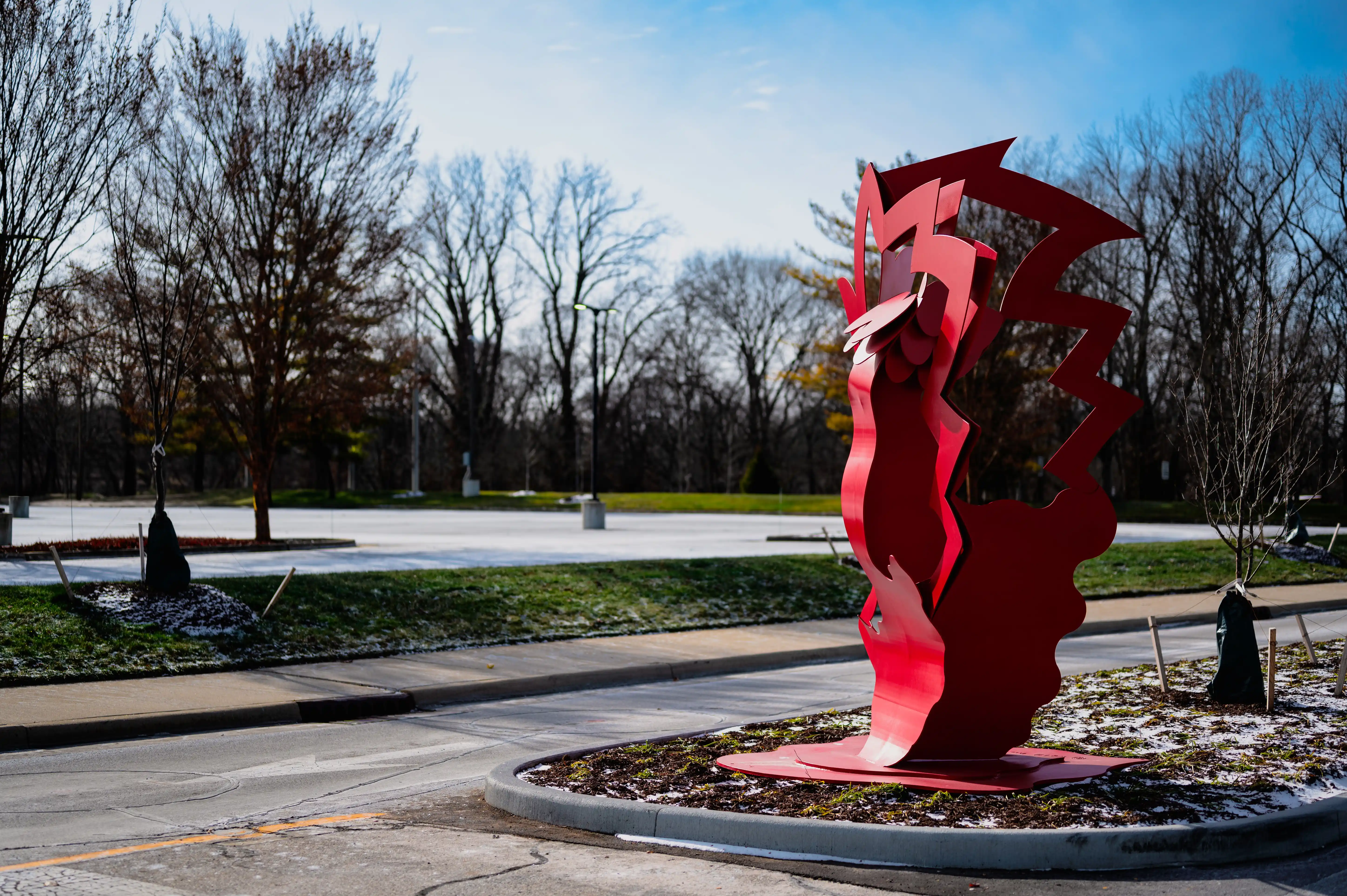 Abstract red sculpture in a park with bare trees and a clear blue sky in the background.