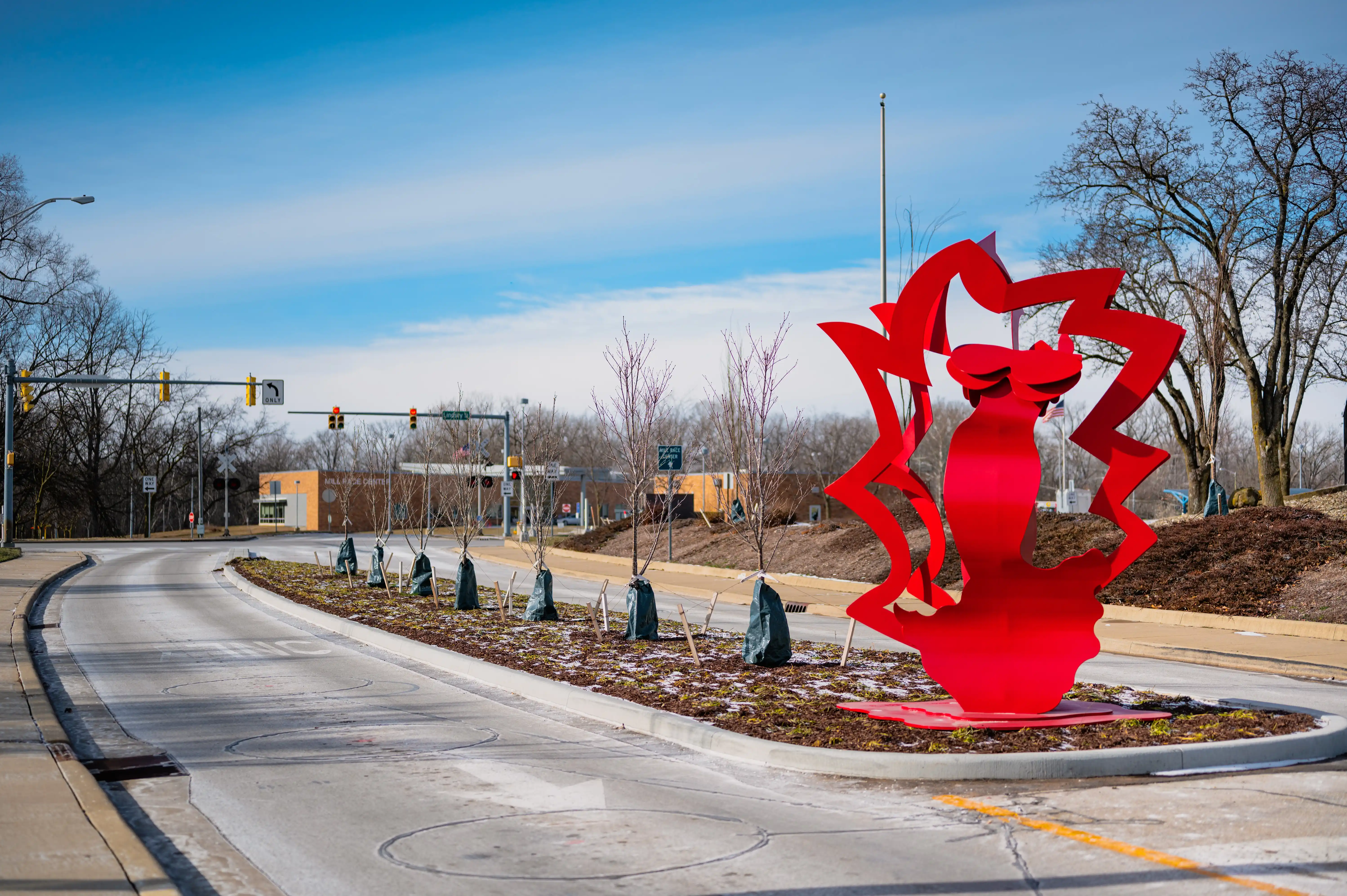 Red abstract sculpture situated in a roundabout with a clear blue sky in the background and bare trees in the distance.