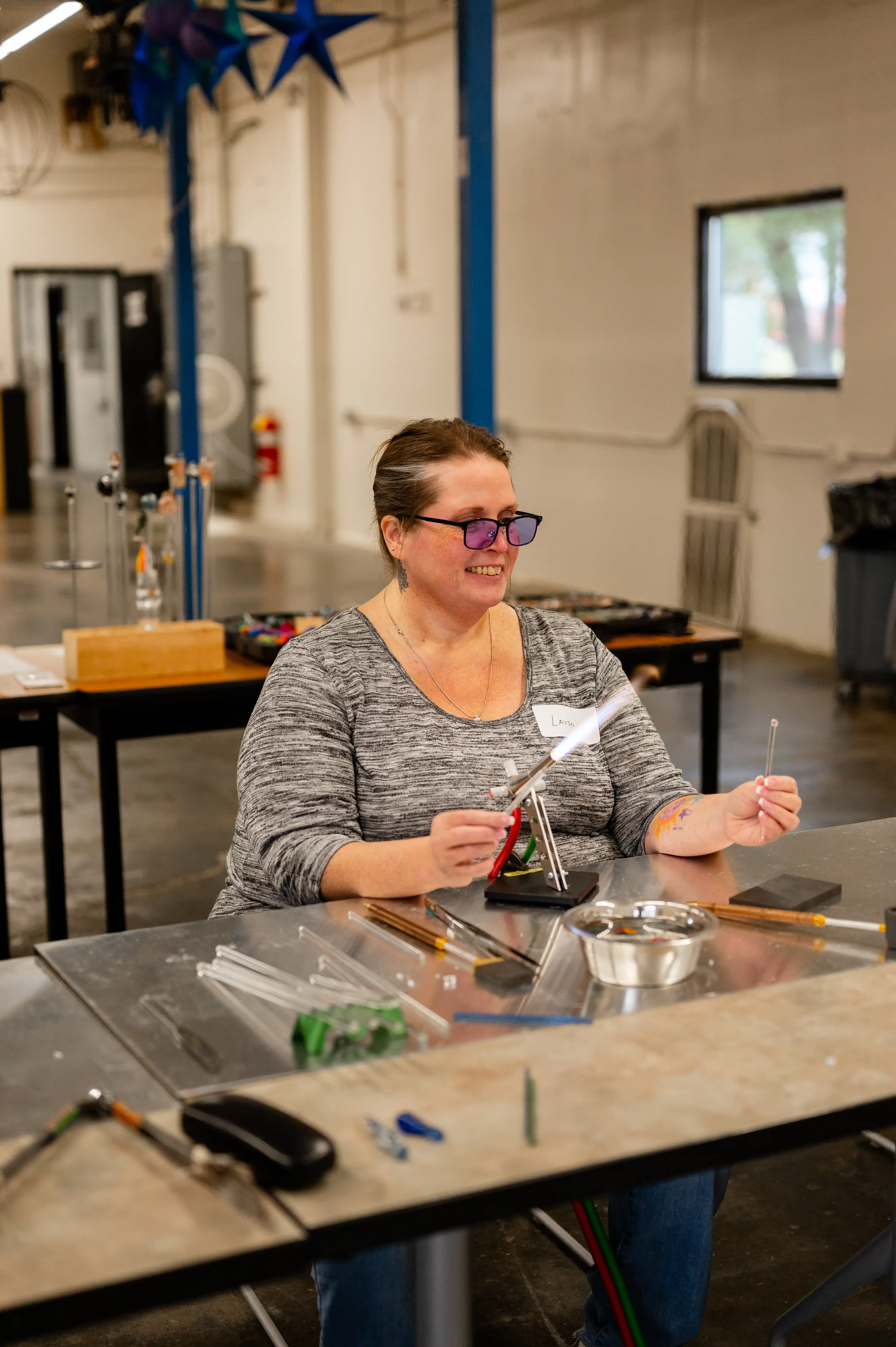 Woman smiling while working with glass rods at a workbench in a workshop setting.