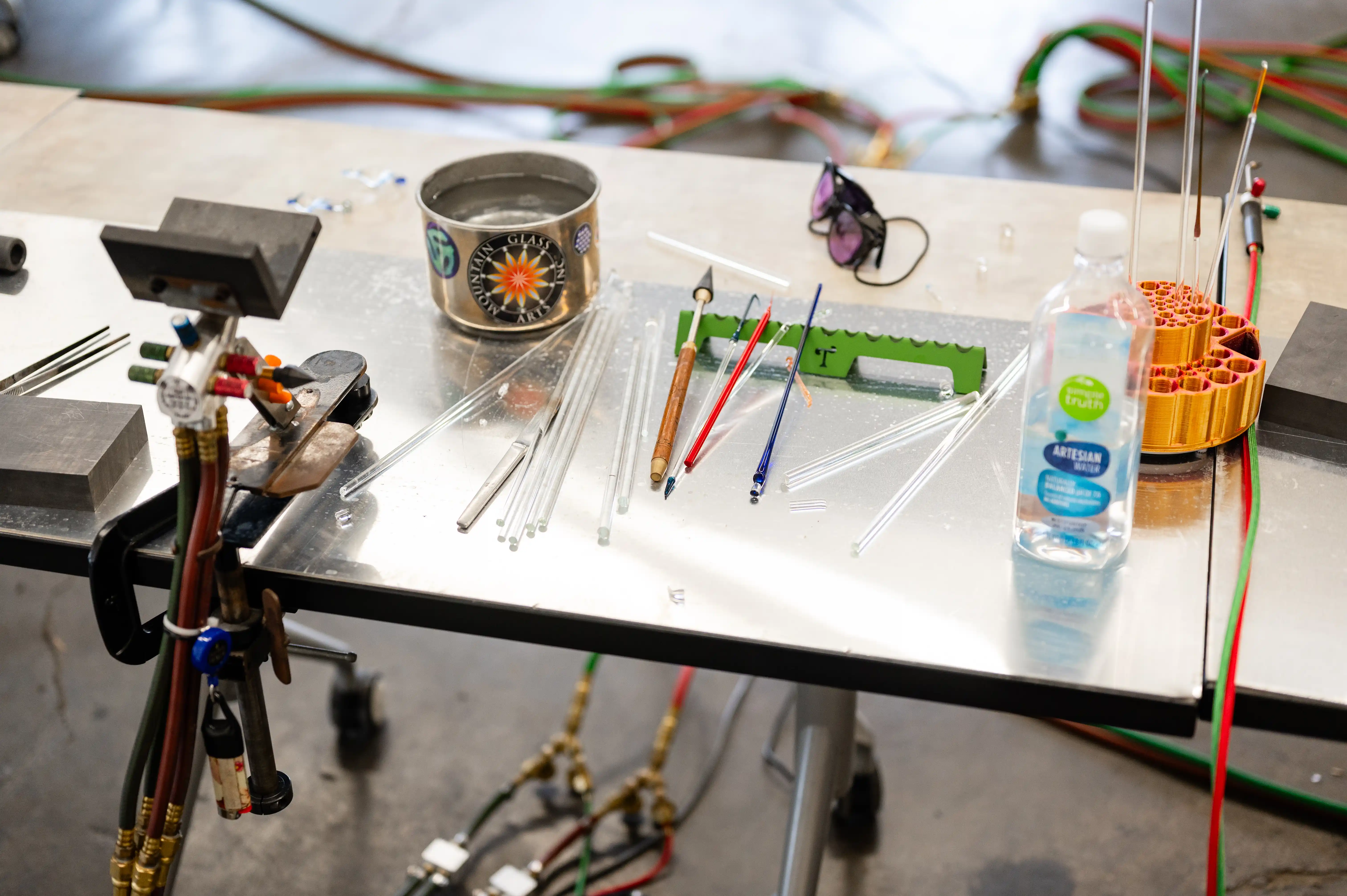 A workbench in a glassblowing studio with various tools, colored glass rods, a tin can, protective glasses, and a bottle of water.