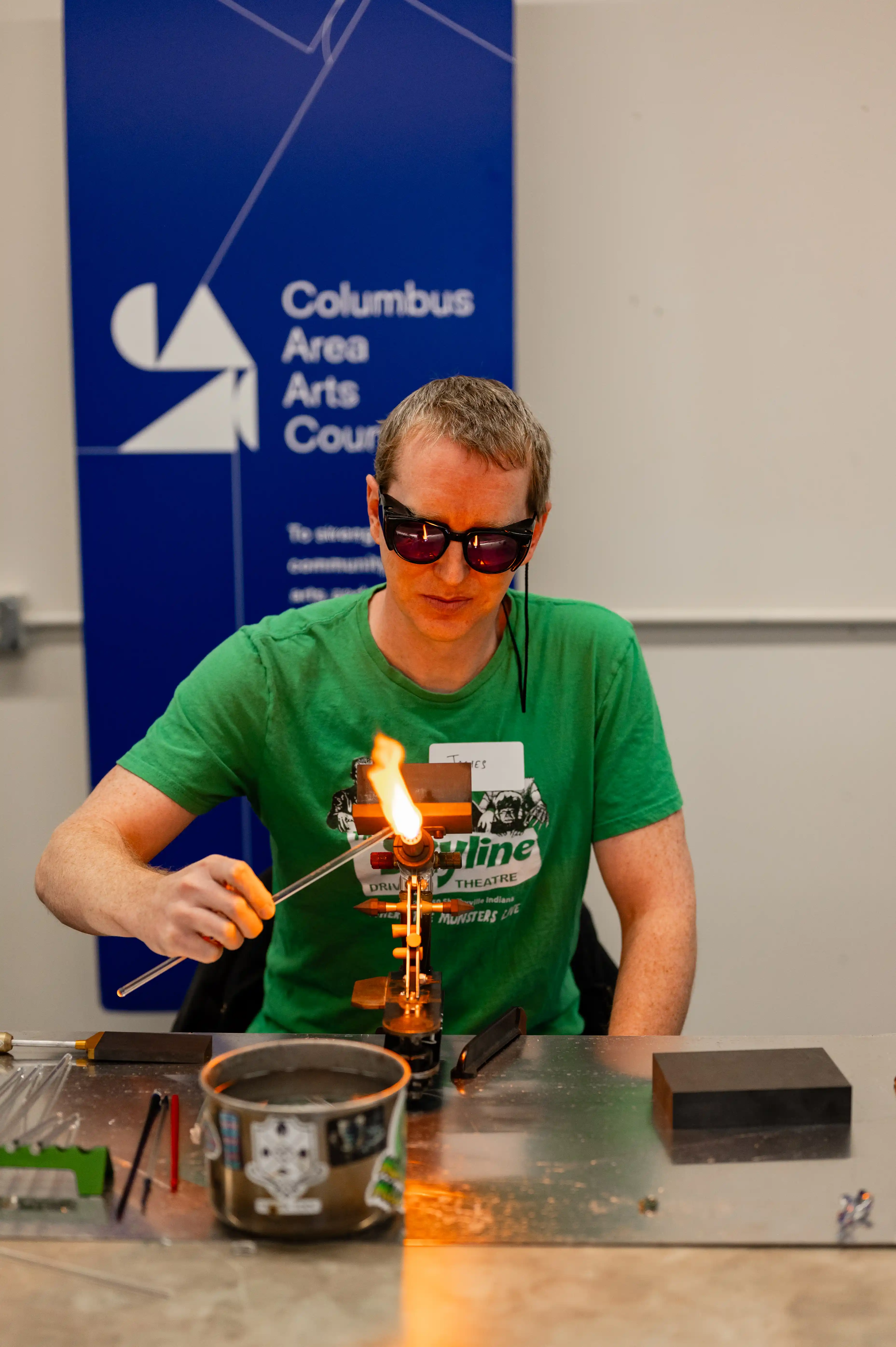 Person wearing protective glasses working with a glass blowing torch at a workshop table with various tools.