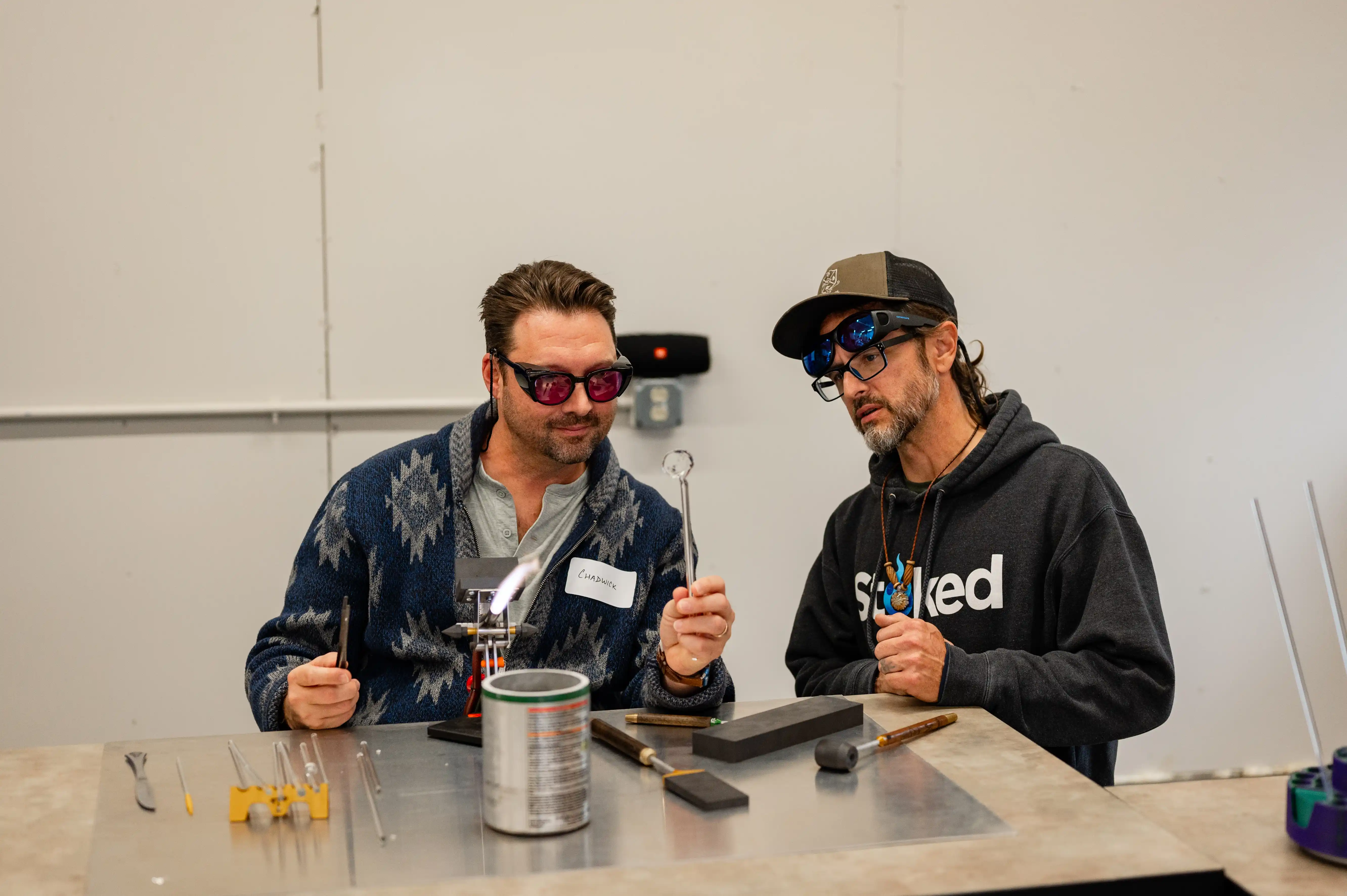 Two people wearing safety glasses working with glassblowing tools at a workshop table.