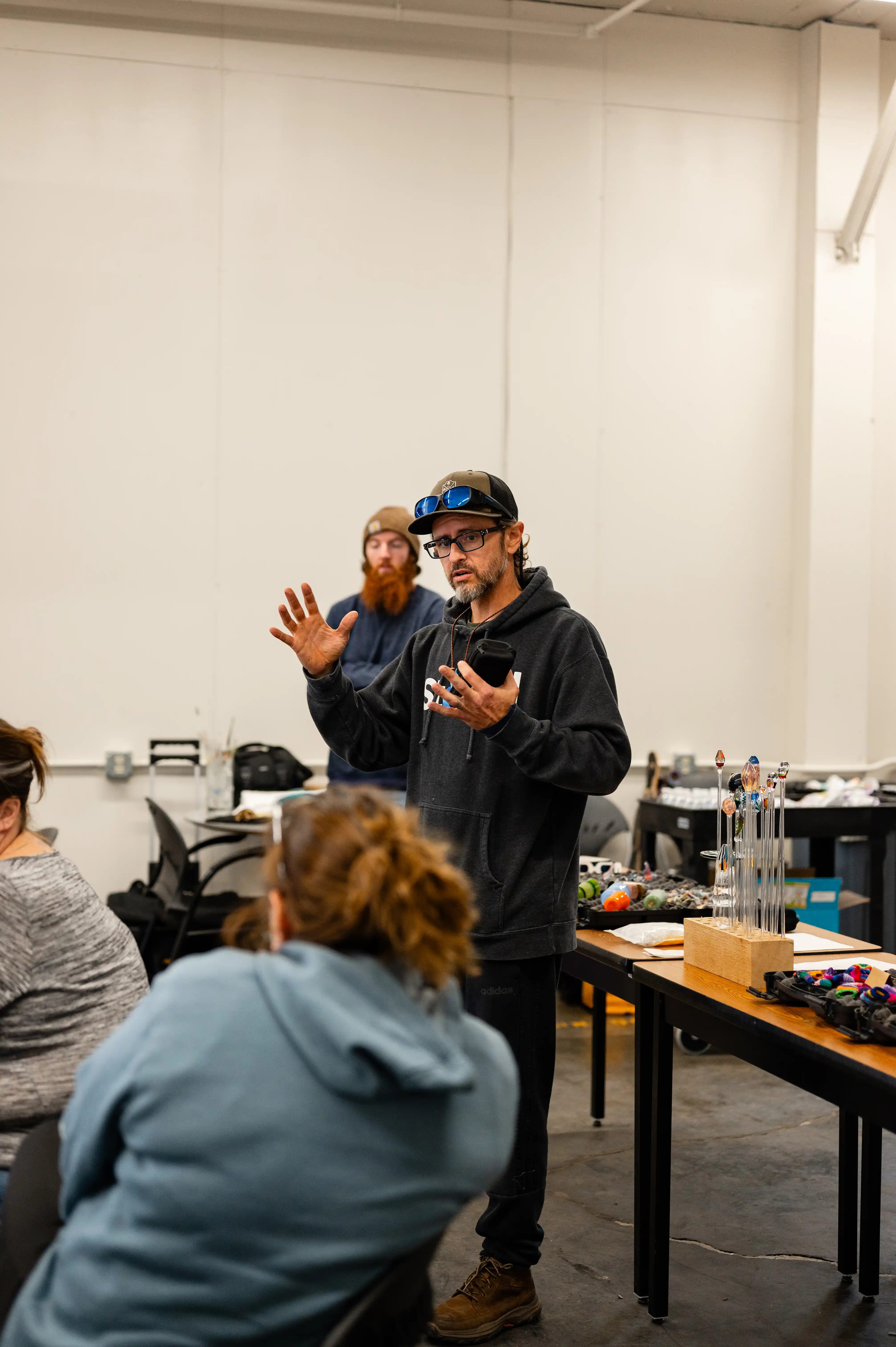 Instructor in glasses and cap gesturing while speaking in a workshop, with attentive students and workbench in foreground.