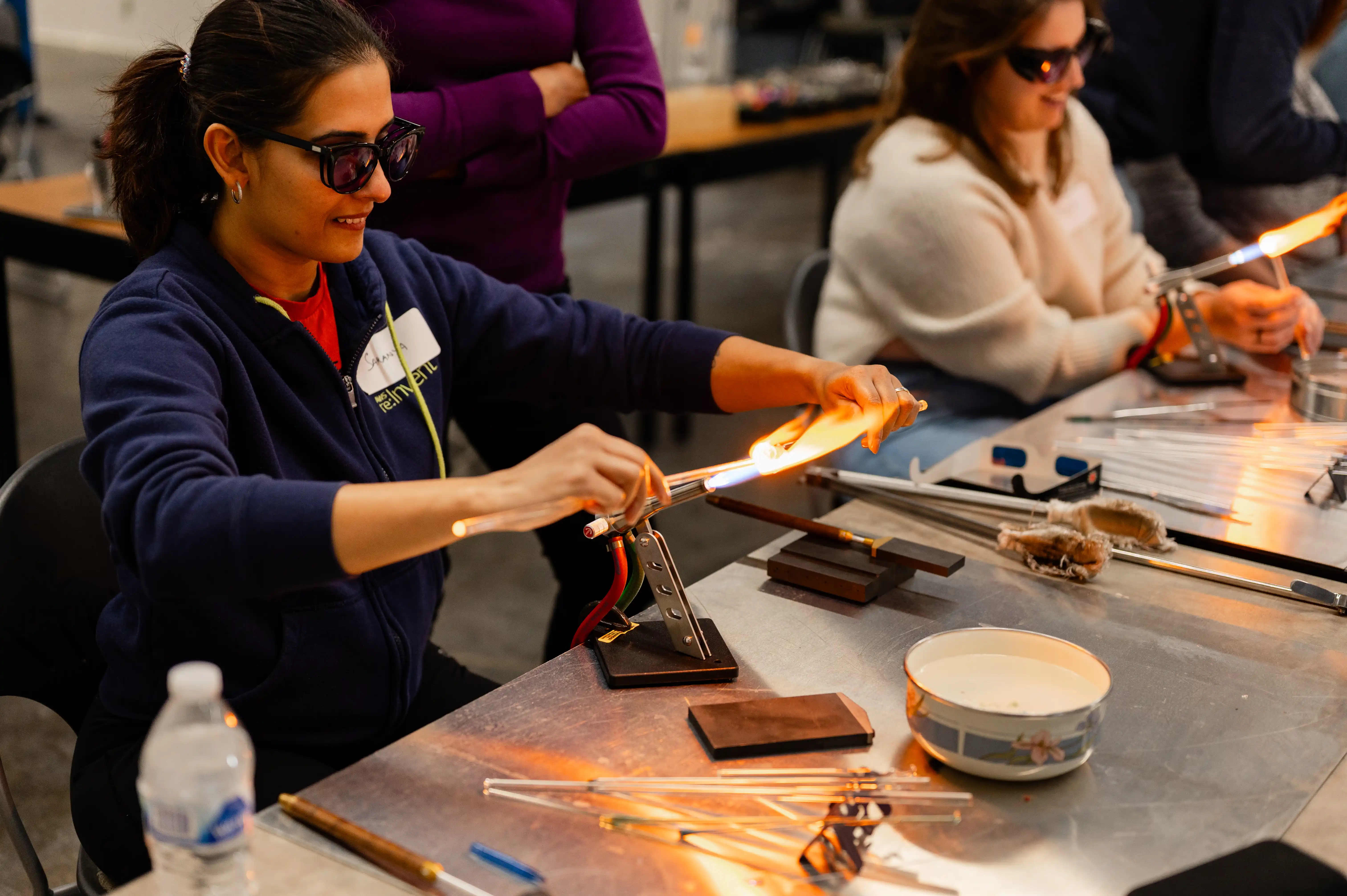 Woman wearing safety glasses shaping glass with a torch in a glassblowing workshop while other participants work in the background.