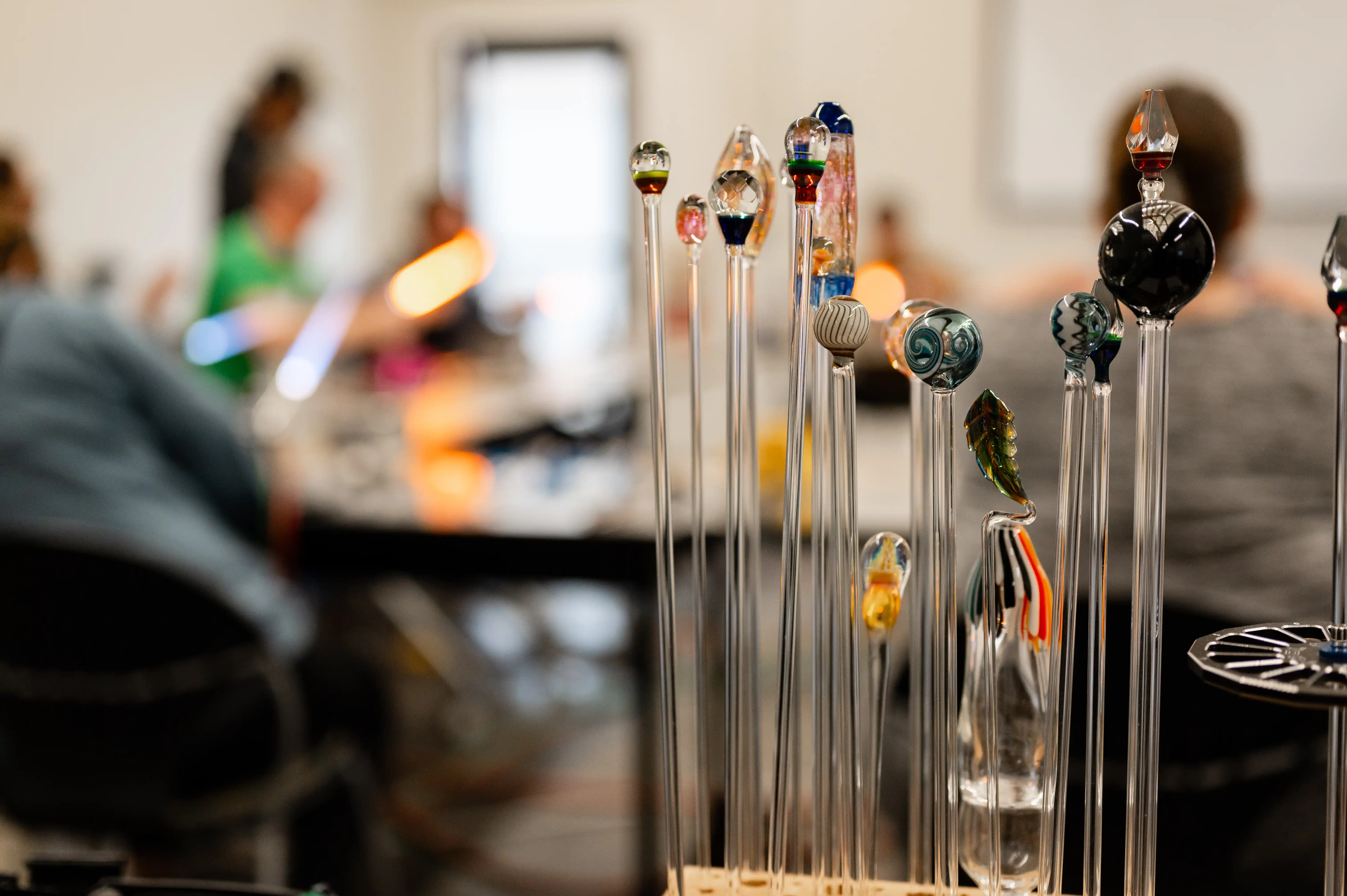 A collection of glass blowing tools with colorful ends on display with a blurred background of a glass blowing workshop and people working.