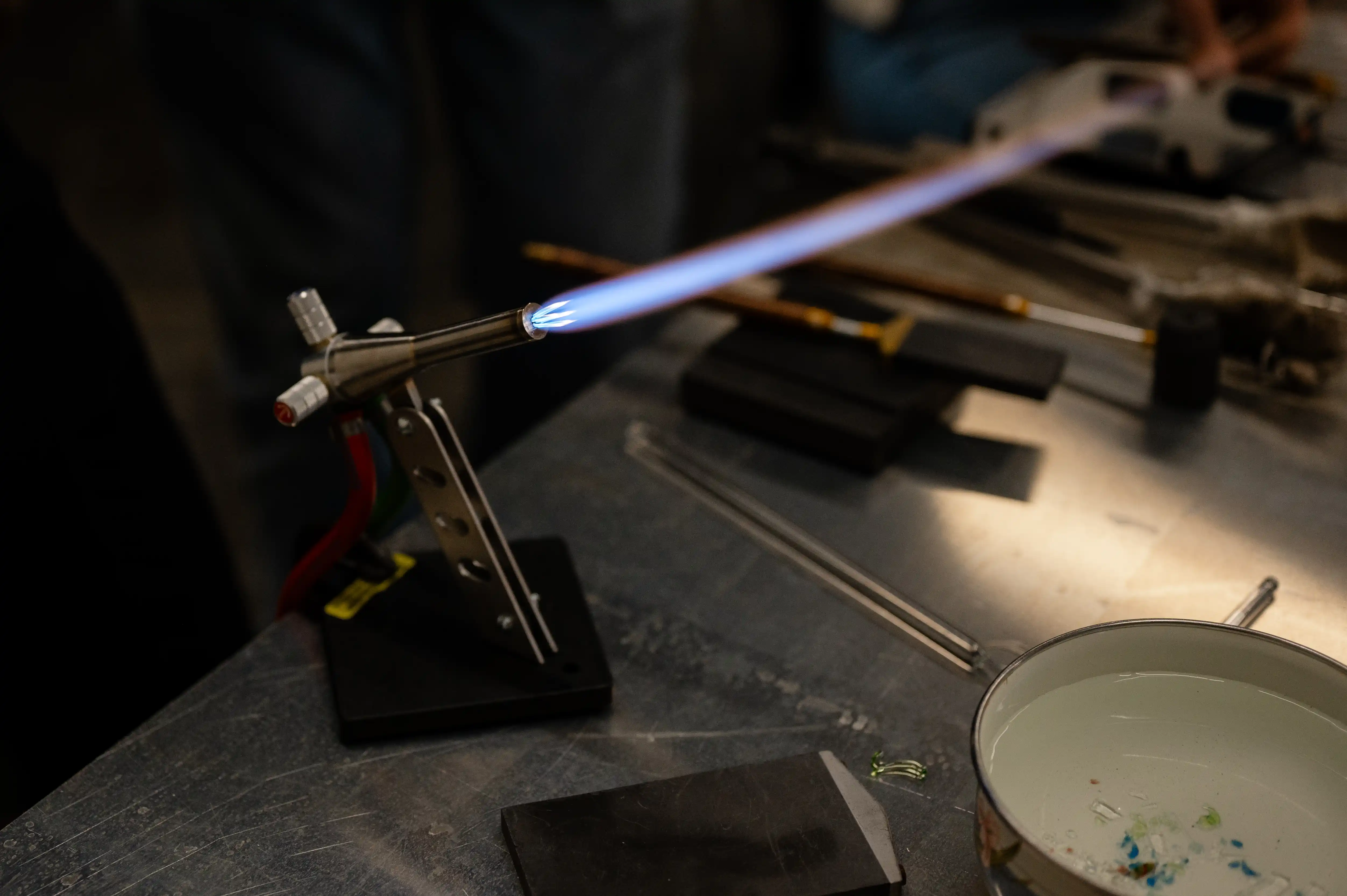 A gas torch with a blue flame mounted on a stand on a workbench, with glassworking tools and materials in the background.